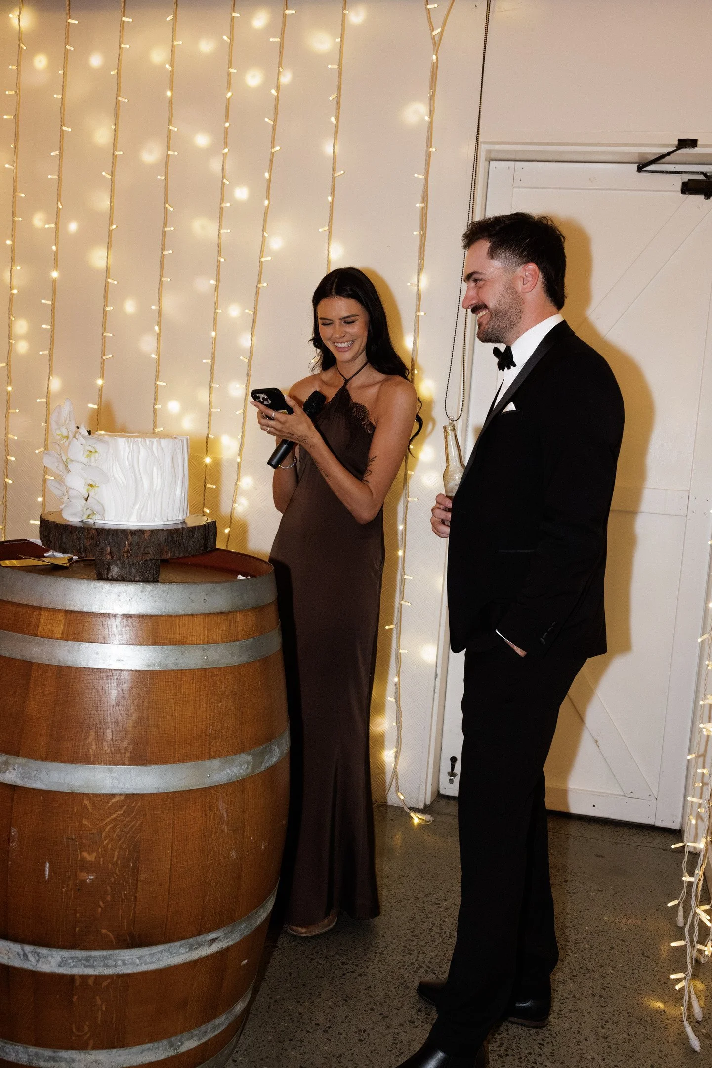 A woman in a brown dress holding a microphone and a phone, smiling while looking at her phone, standing next to a man in a black tuxedo holding a drink, at a celebration with string lights and a cake on a barrel in the background.