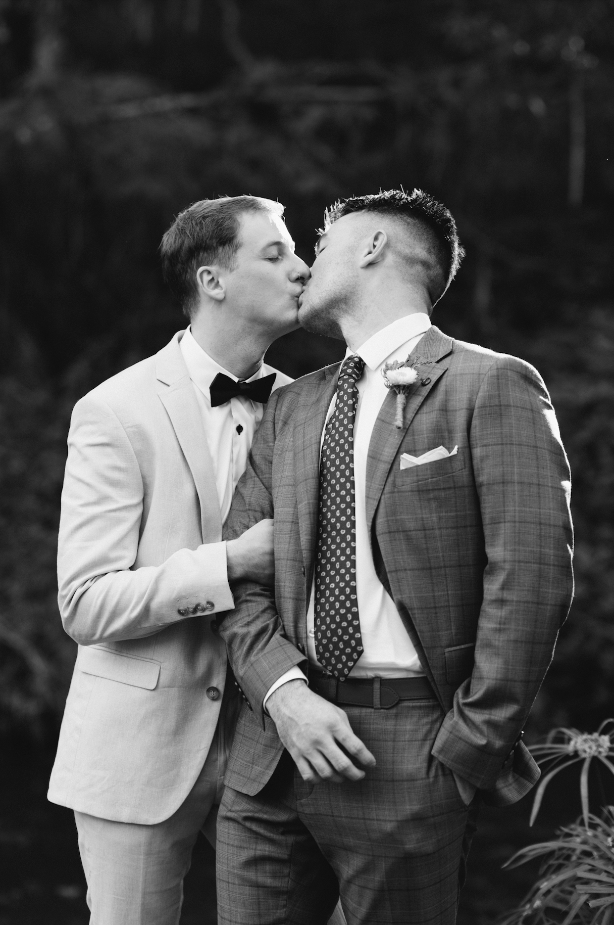 Two men in suits sharing a kiss outdoors, one wearing a light-colored suit with a bow tie and the other a darker plaid suit with a patterned tie.