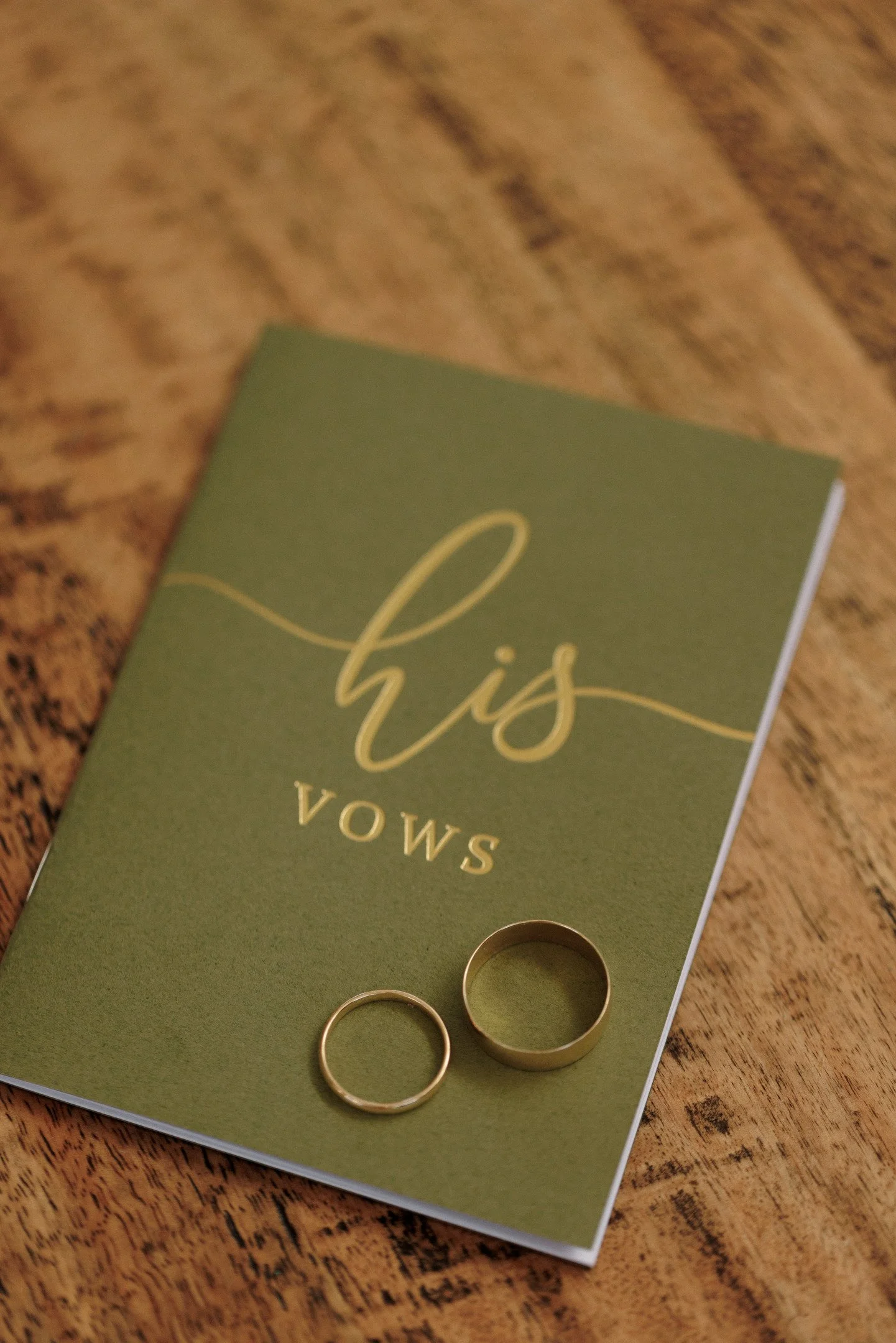 Two gold wedding bands placed on a green card that has the word "Vows" written on it, with a wooden surface in the background.