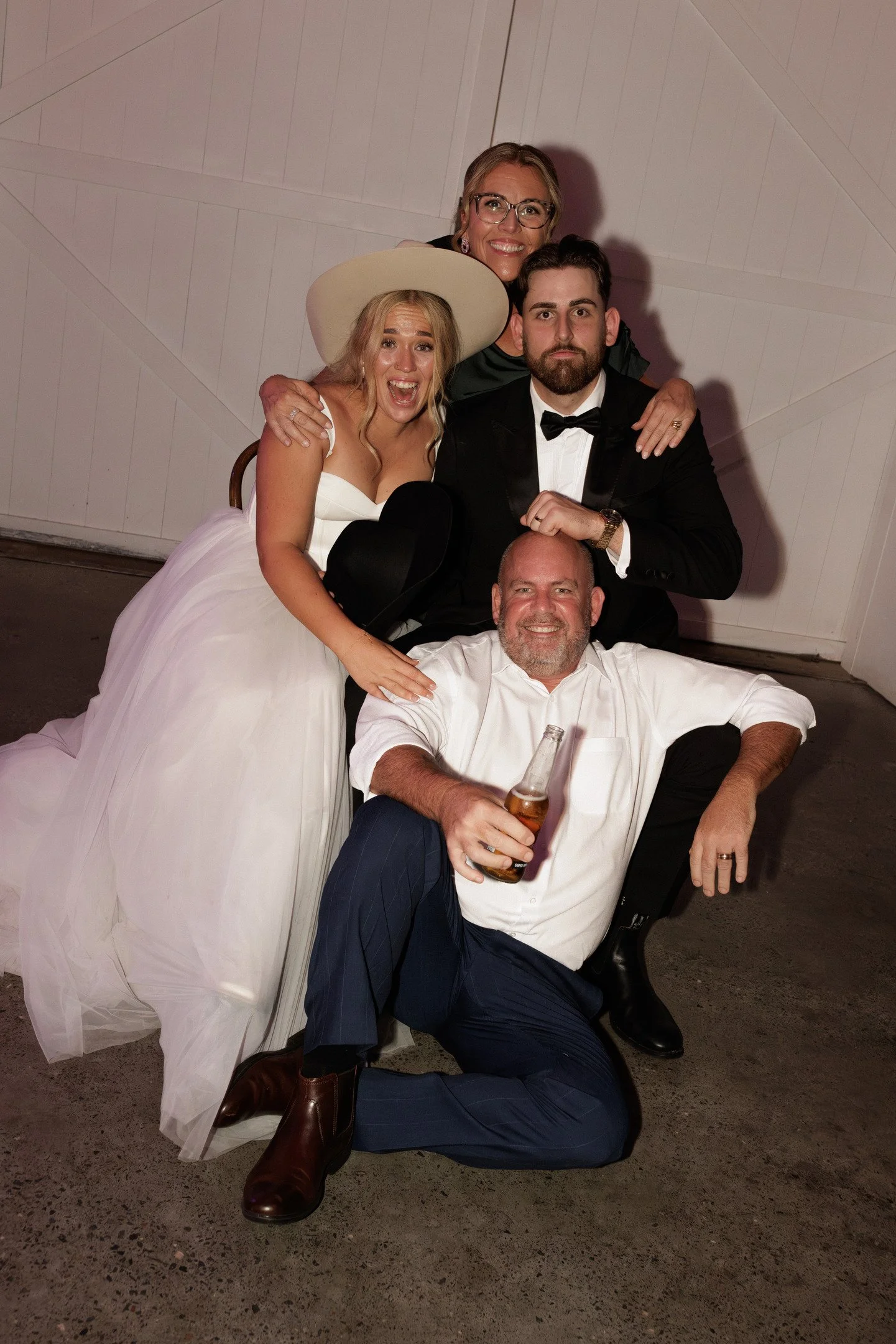 A group of four people at a celebration or wedding, with one woman in a white dress and large hat, another woman with glasses and earrings, a man in a tuxedo, and another man holding a beer bottle, all smiling or making funny faces, in front of a whi