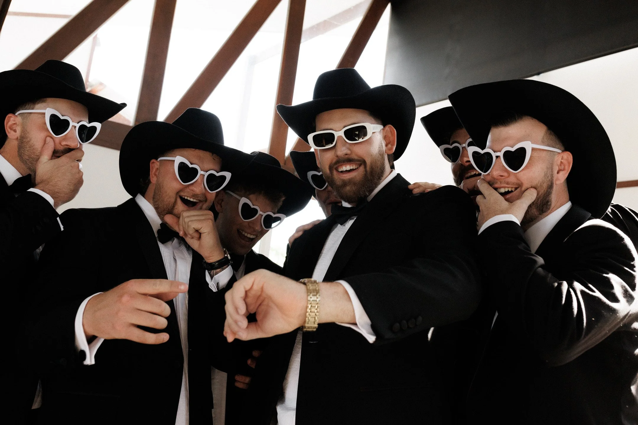 Group of men dressed in tuxedos, wearing large black hats and heart-shaped sunglasses, smiling and laughing together at a celebration.