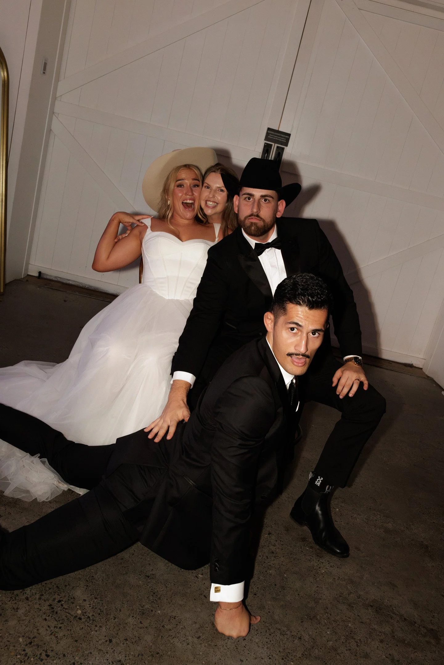 Group of five friends dressed in formal attire, with a woman in a white wedding dress and hat, and four men in tuxedos and hats, posing in front of a white wall with a wooden door.