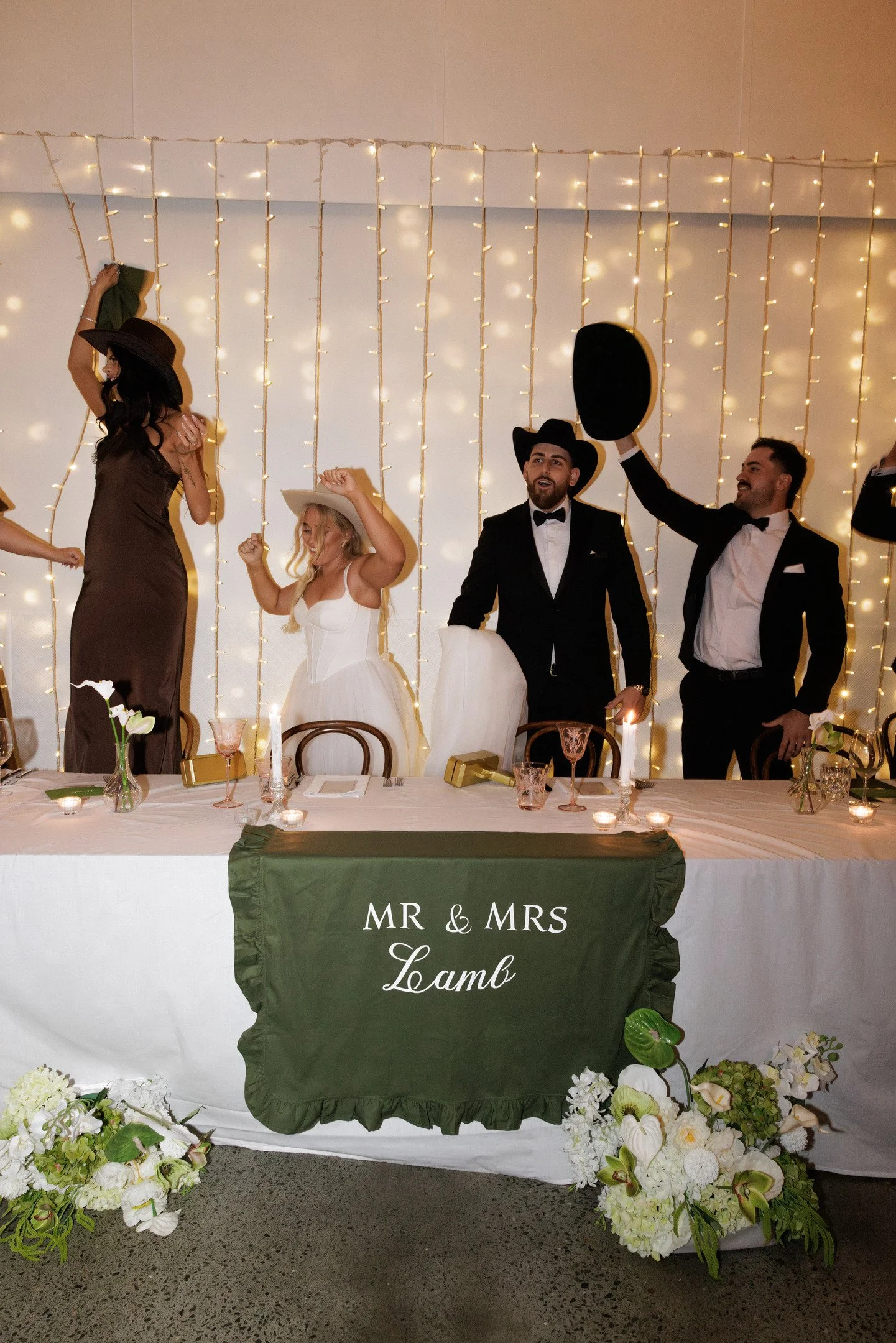 Wedding reception table with a green cloth that reads "MR & MRS Lamb." The table has white flowers, candles, and wine glasses. Behind the table, people are dancing, wearing formal and cowboy hats, with string lights on the wall.