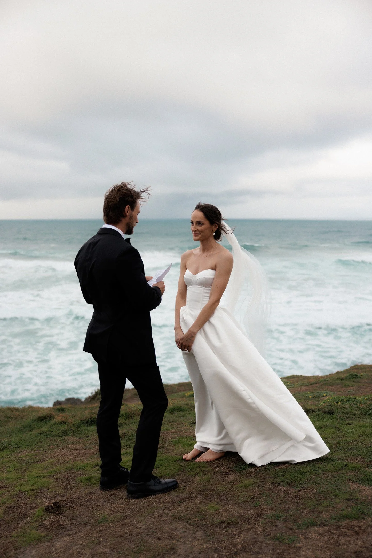 A man in a black tuxedo is holding a paper, standing on a grassy area by the ocean, facing a woman in a white wedding dress with a veil, with waves behind them.