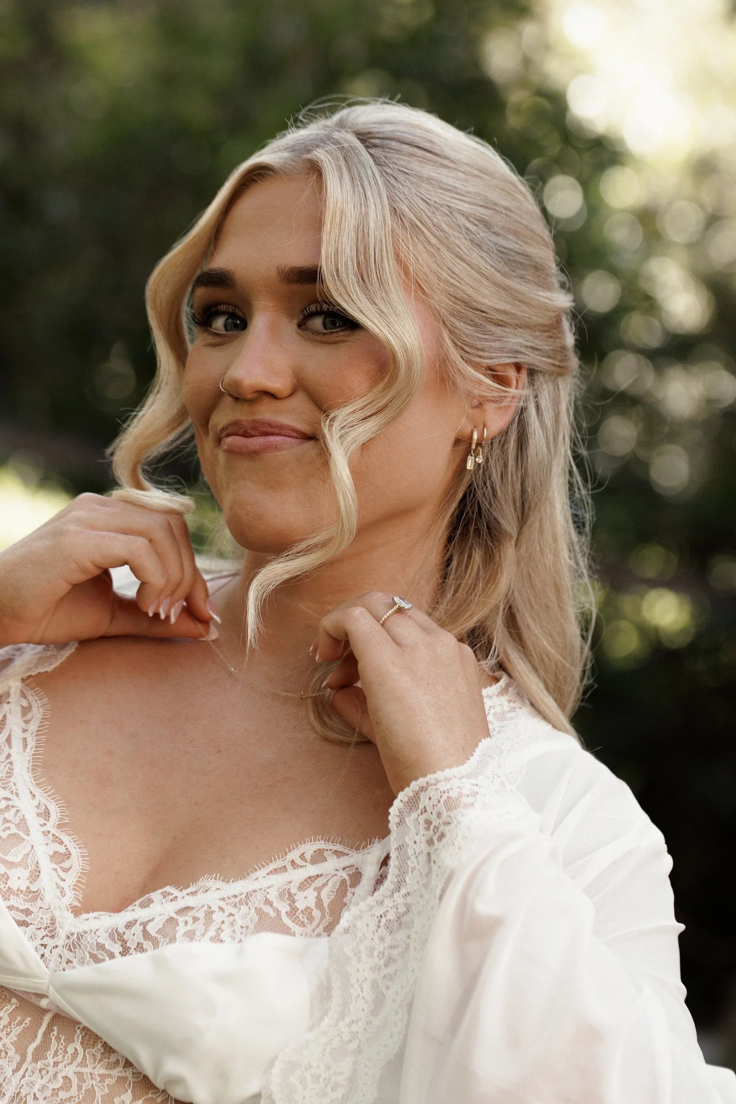A woman with blonde wavy hair and blue eyes wearing a white lace blouse and jewelry, outdoors with blurred green trees in the background.