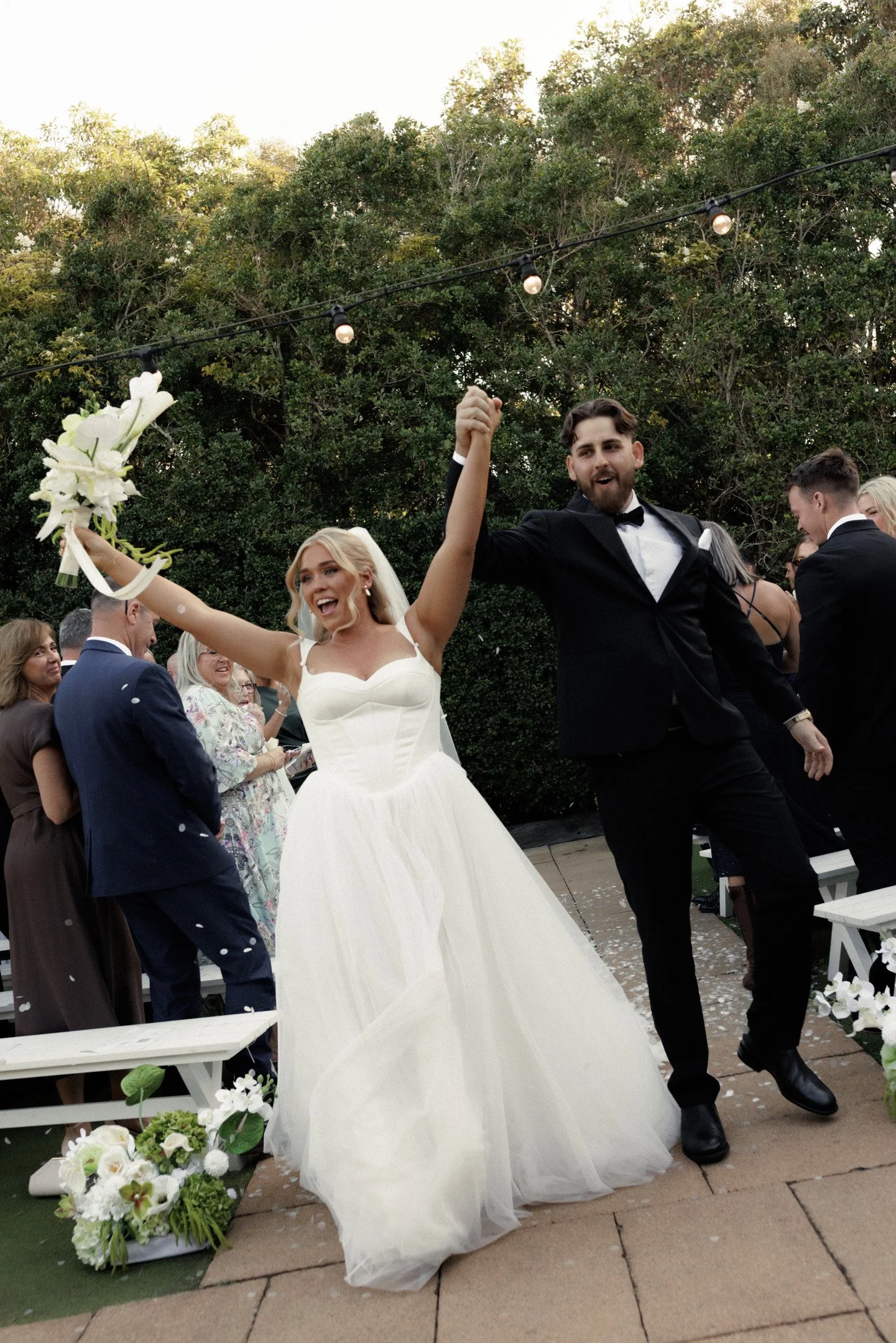 Celebrating wedding couple outdoors with guests, bride in white gown holding bouquet, groom in black tuxedo, surrounded by greenery and string lights.