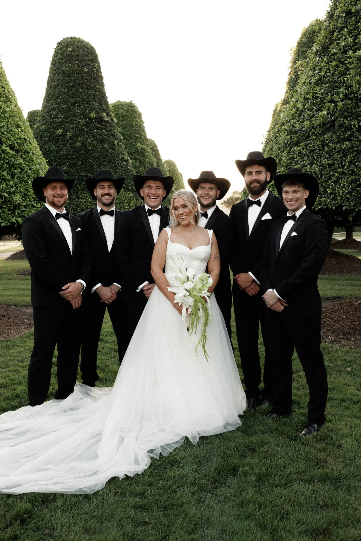 A bride in a white wedding dress and bouquet standing with six groomsmen dressed in black tuxedos and black cowboy hats, outdoors among green trees.