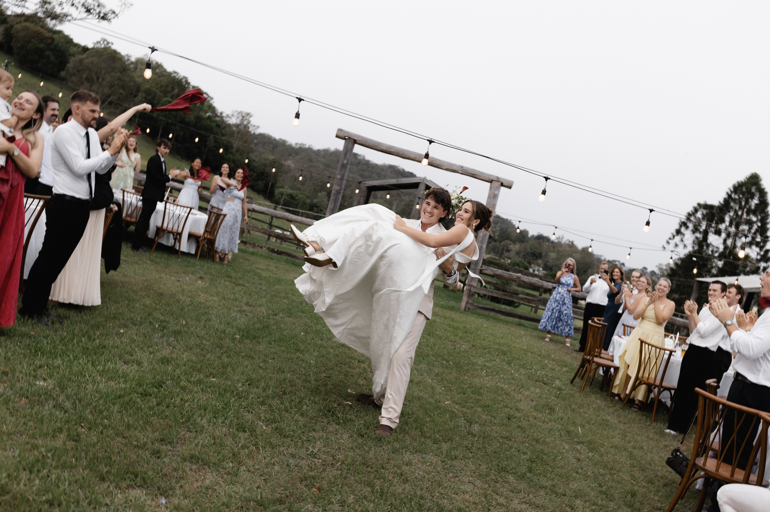 A man in a beige suit is carrying a smiling woman in a white dress during an outdoor wedding reception, surrounded by guests clapping and celebrating under string lights on a grassy field.