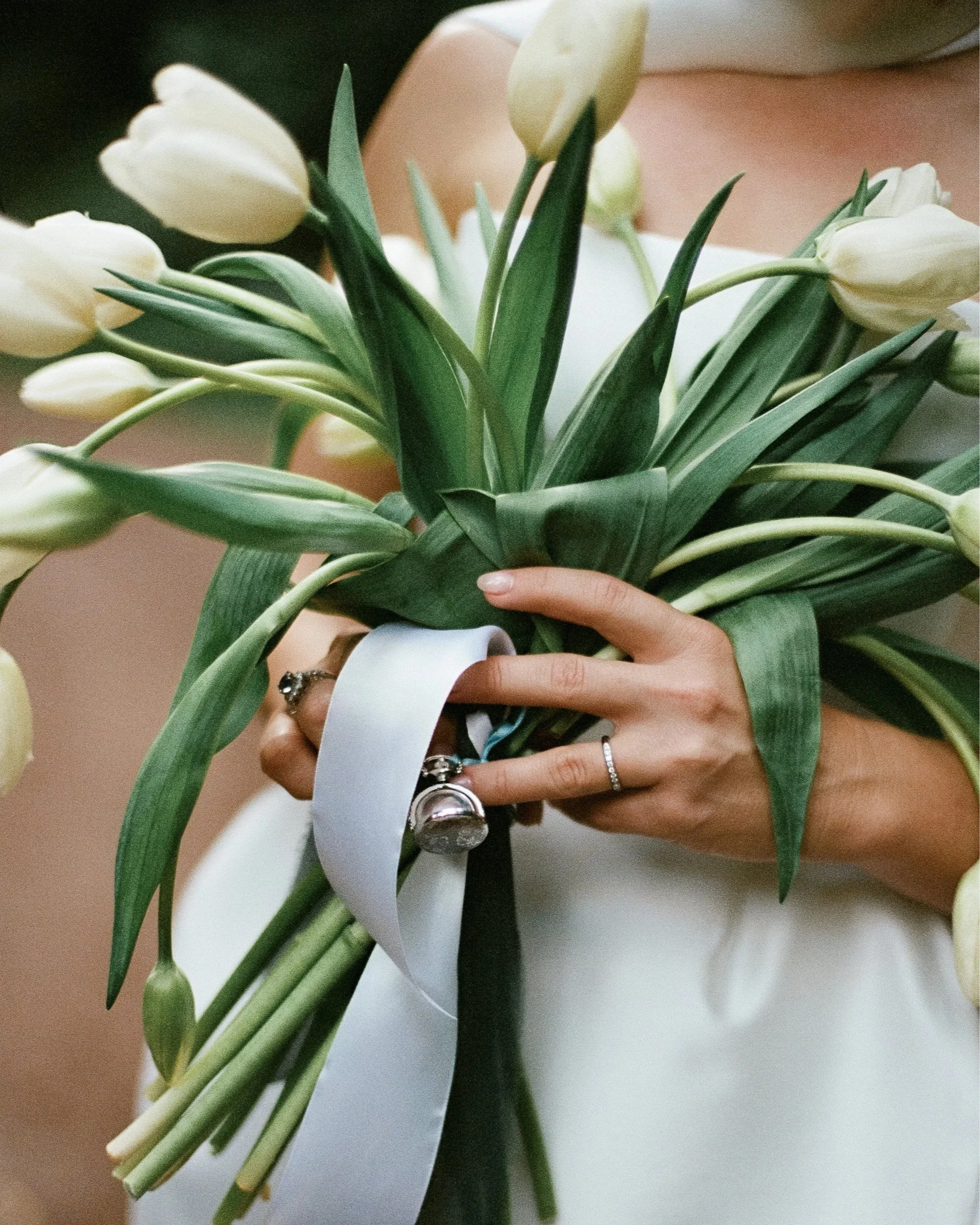 Person holding a bouquet of white tulips with green leaves, decorated with white and black ribbons, wearing rings.