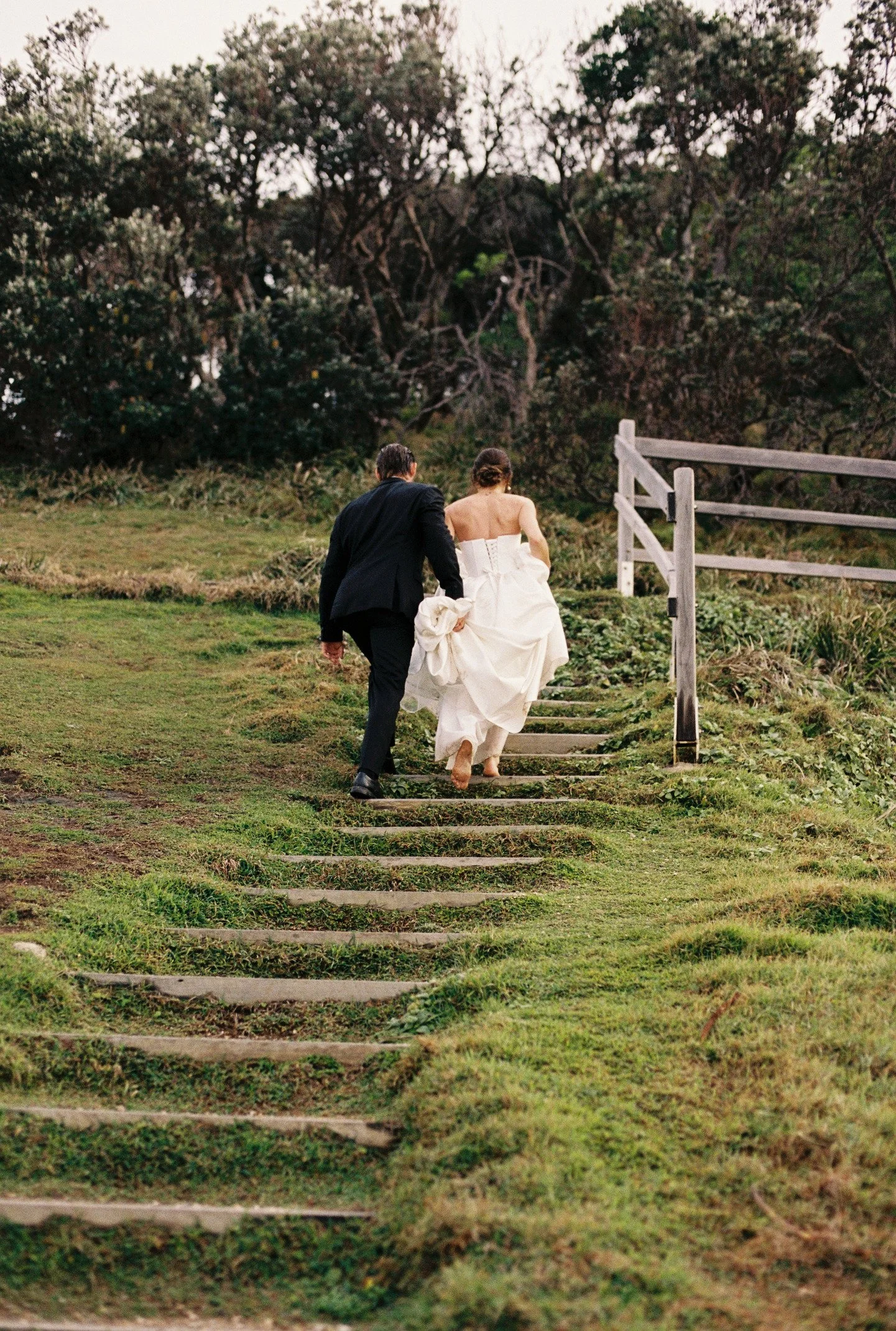A man in a black suit and a woman in a white wedding gown walking up outdoor wooden stairs on a grassy area, surrounded by trees.