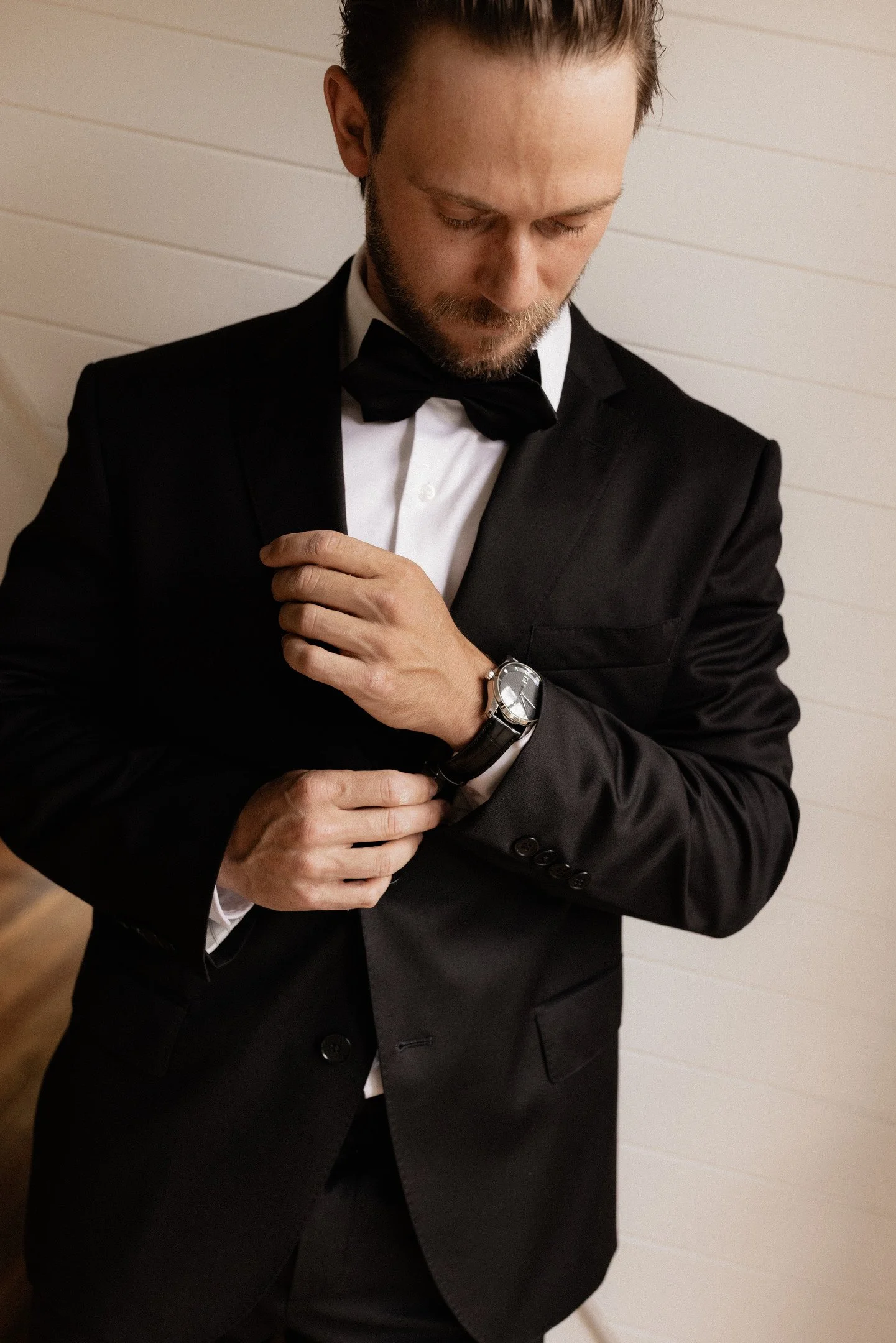 A man wearing a black tuxedo and bow tie is adjusting his watch. He has short, dark hair and a beard, standing against a light-colored wall.