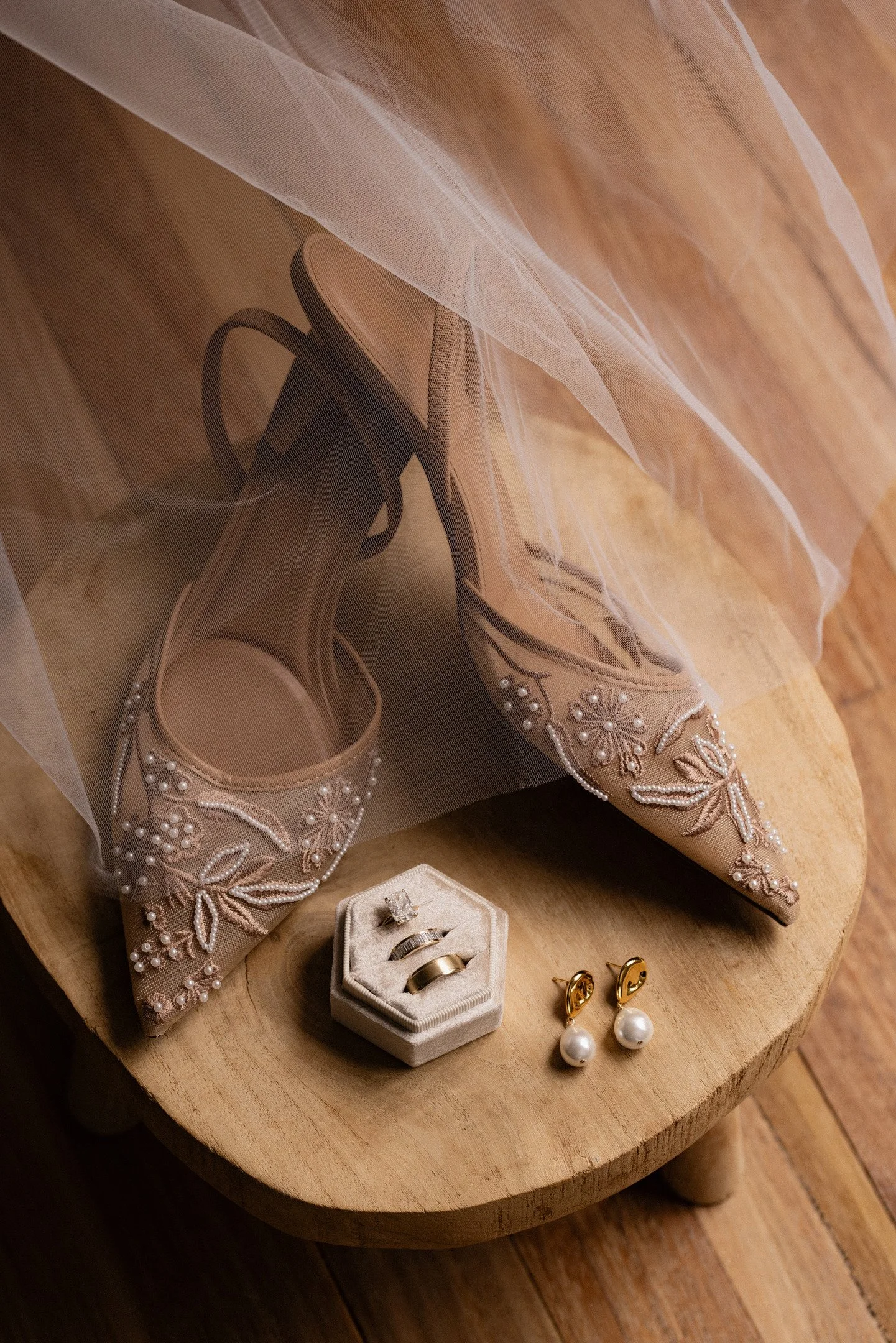 A pair of beige bridal shoes with floral embroidery and pearl embellishments, a ring set in a gray jewelry box, and pearl and gold earrings on a round wooden table.