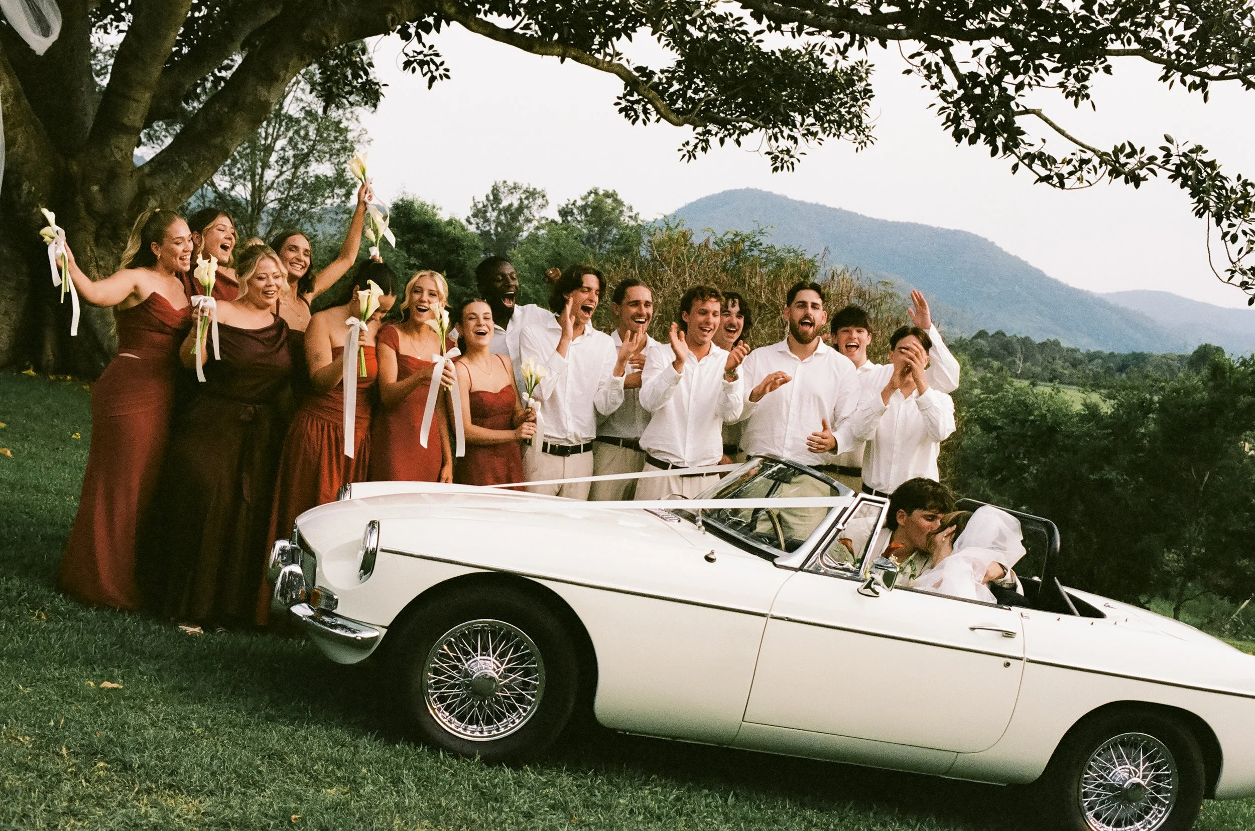Group of joyful people celebrating around a vintage white convertible car outdoors, with mountains and trees in the background. The group includes men and women, some holding flowers, and a couple in the car appears to be kissing, suggesting a weddin