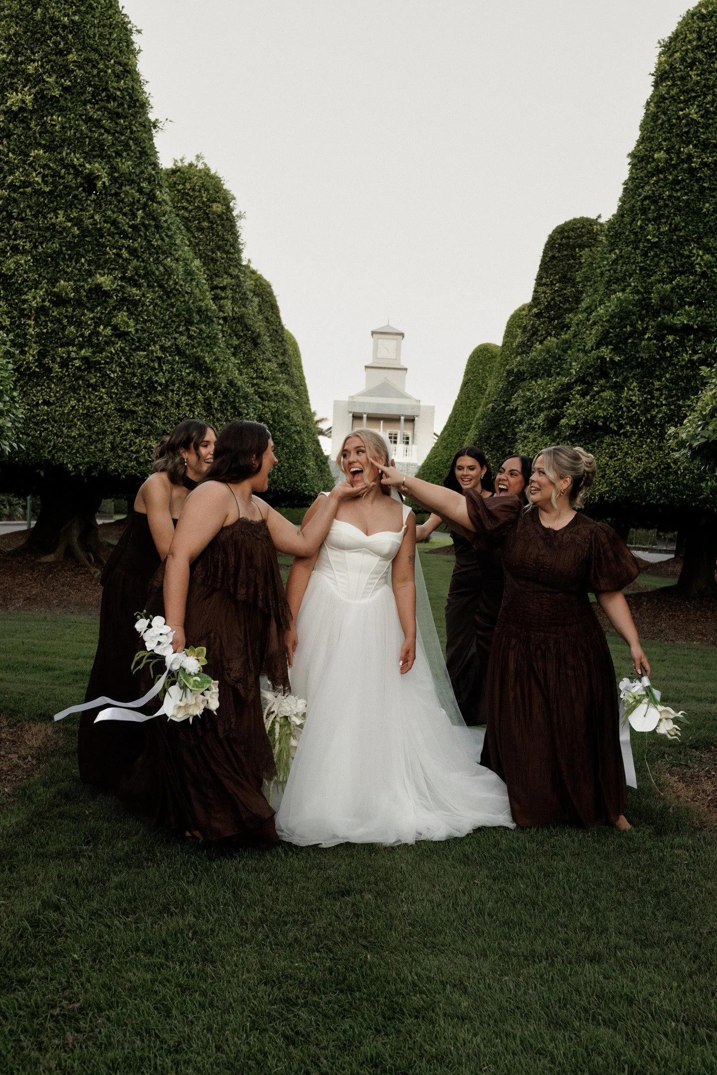 Group of women at a wedding, with one bride in a white gown, surrounded by bridesmaids in dark brown dresses, in a garden with tall, trimmed trees, and a white building with a clock tower in the background.
