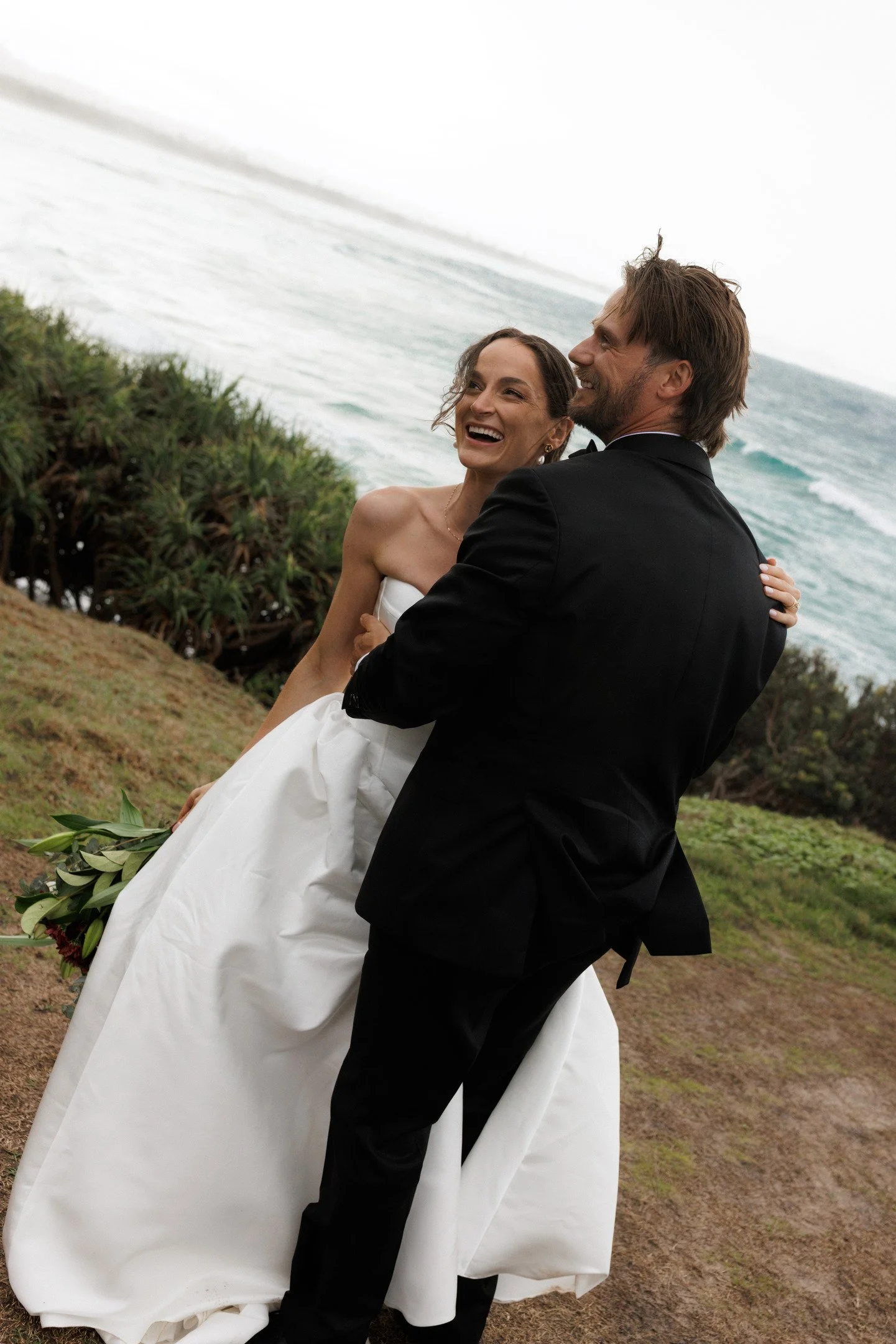 A smiling bride and groom embracing outdoors near the ocean, with greenery and waves in the background, during their wedding ceremony.