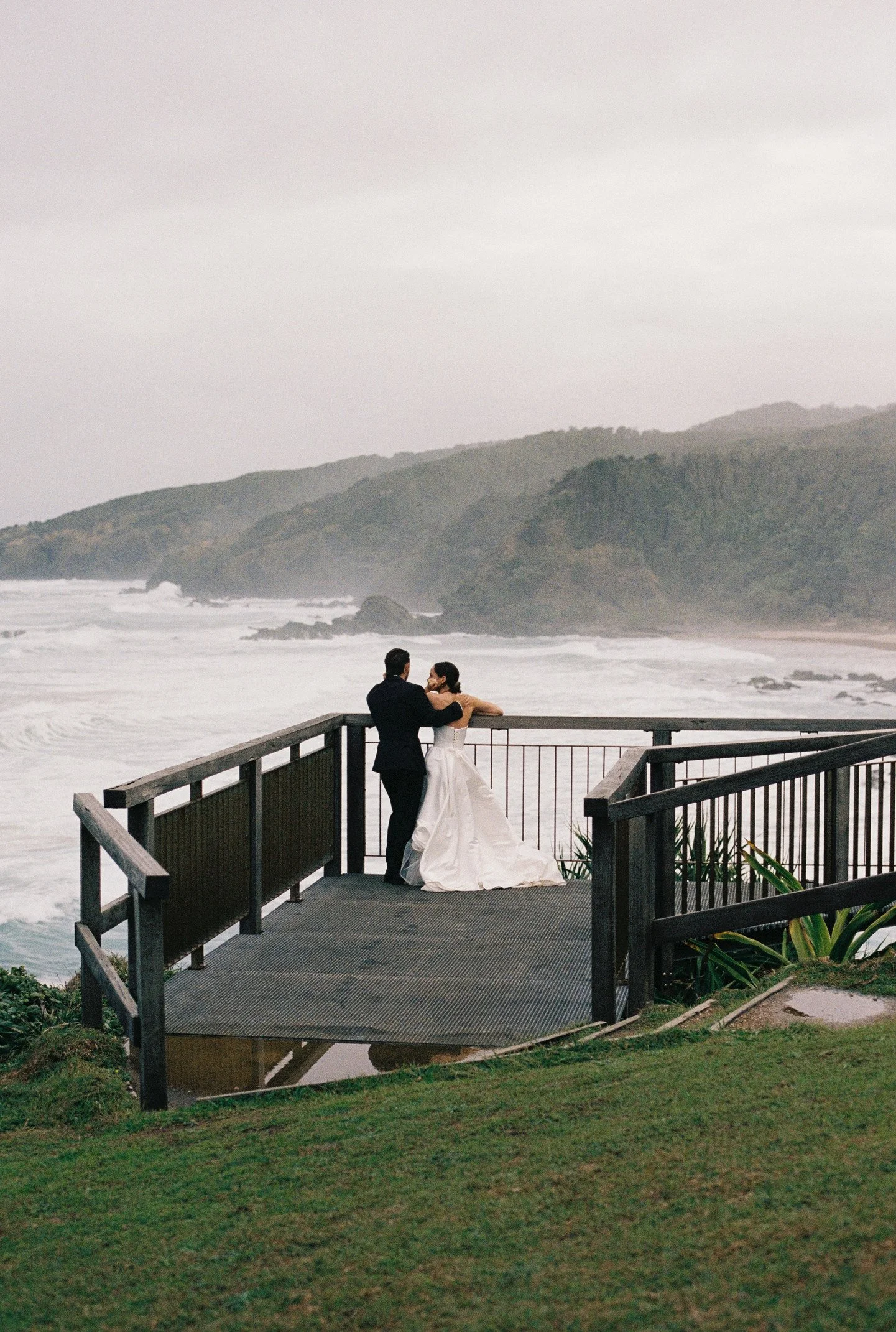 A couple in wedding attire standing on a wooden deck overlooking the ocean with foggy mountains in the background.