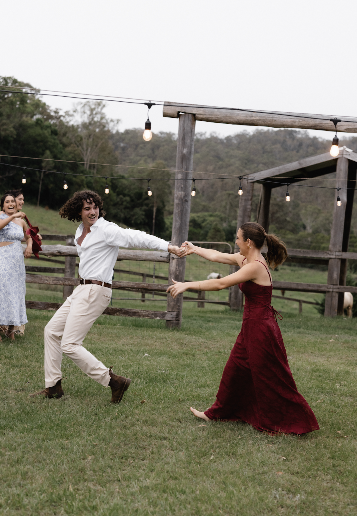 People dancing and having fun outdoors on a farm with string lights, fence, and trees in the background.
