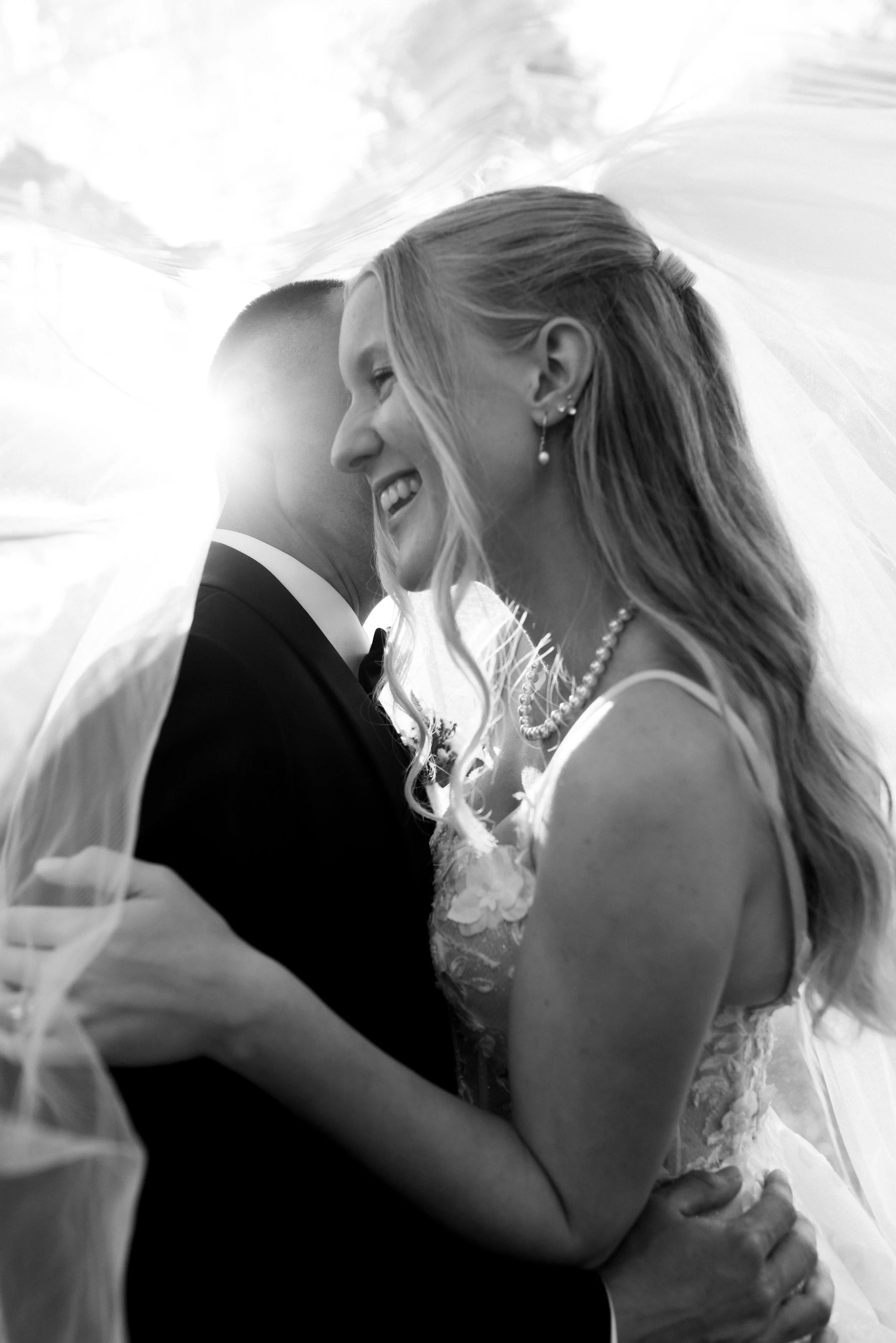A black-and-white photograph of a smiling bride and groom embracing each other closely, with the bride wearing pearl jewelry and a wedding dress, and the groom in a tuxedo.