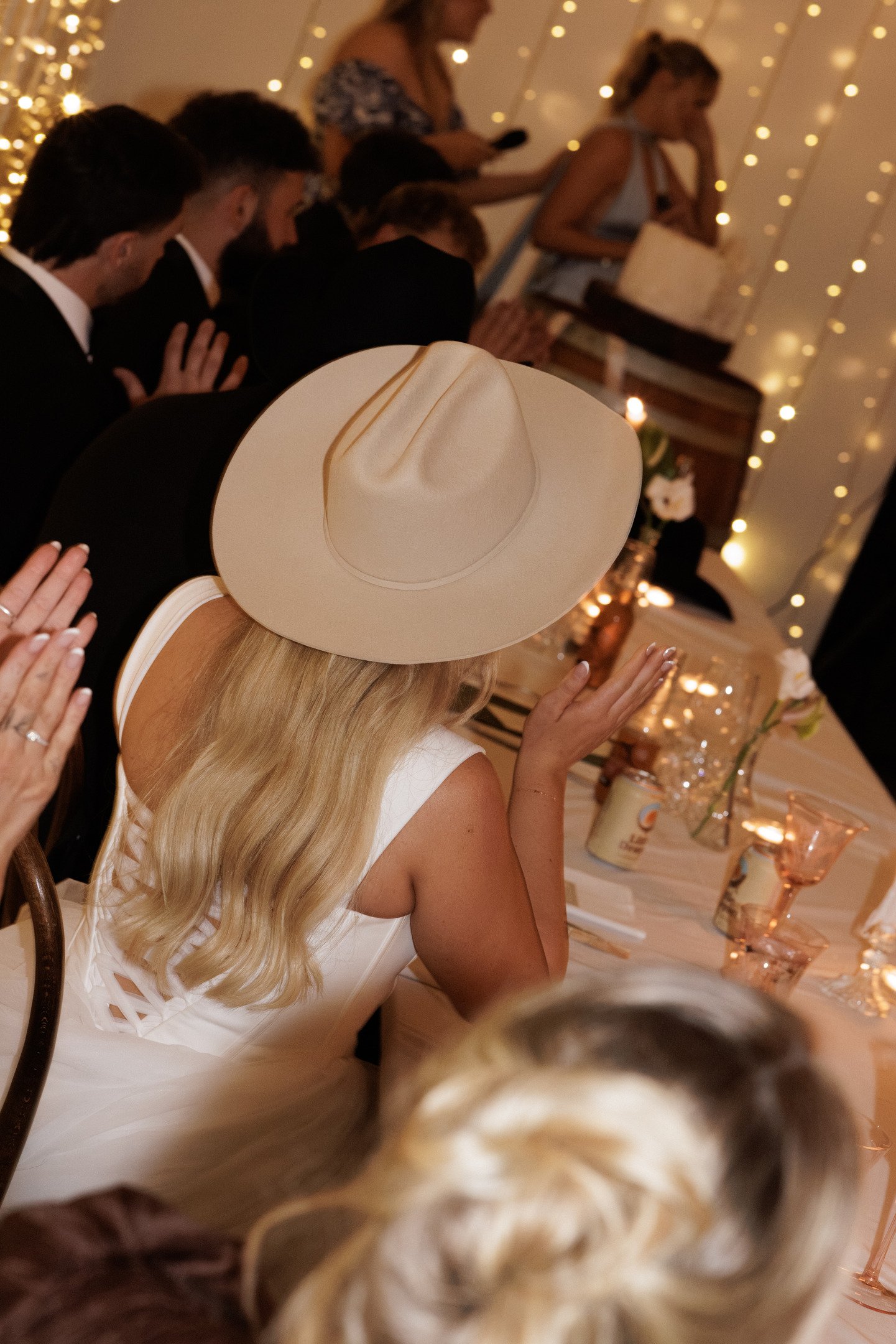 People participating in a prayer or blessing ceremony at a formal event, with some hands raised and a woman in a large cream-colored hat in the foreground.