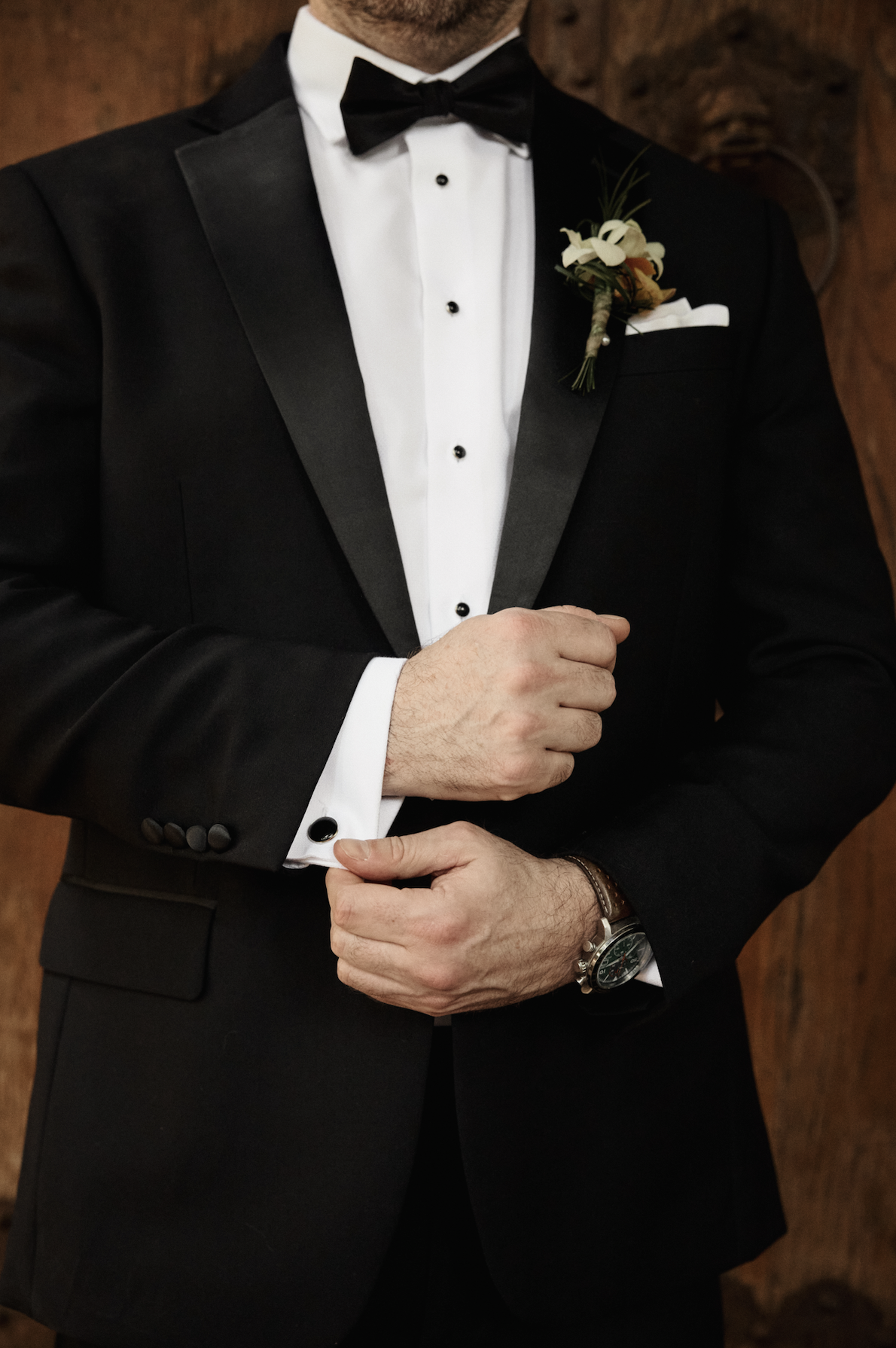 Close-up of a man in a black tuxedo with a white dress shirt and black bow tie, adjusting his cufflink, with a boutonniere on his lapel and a wristwatch on his left wrist.