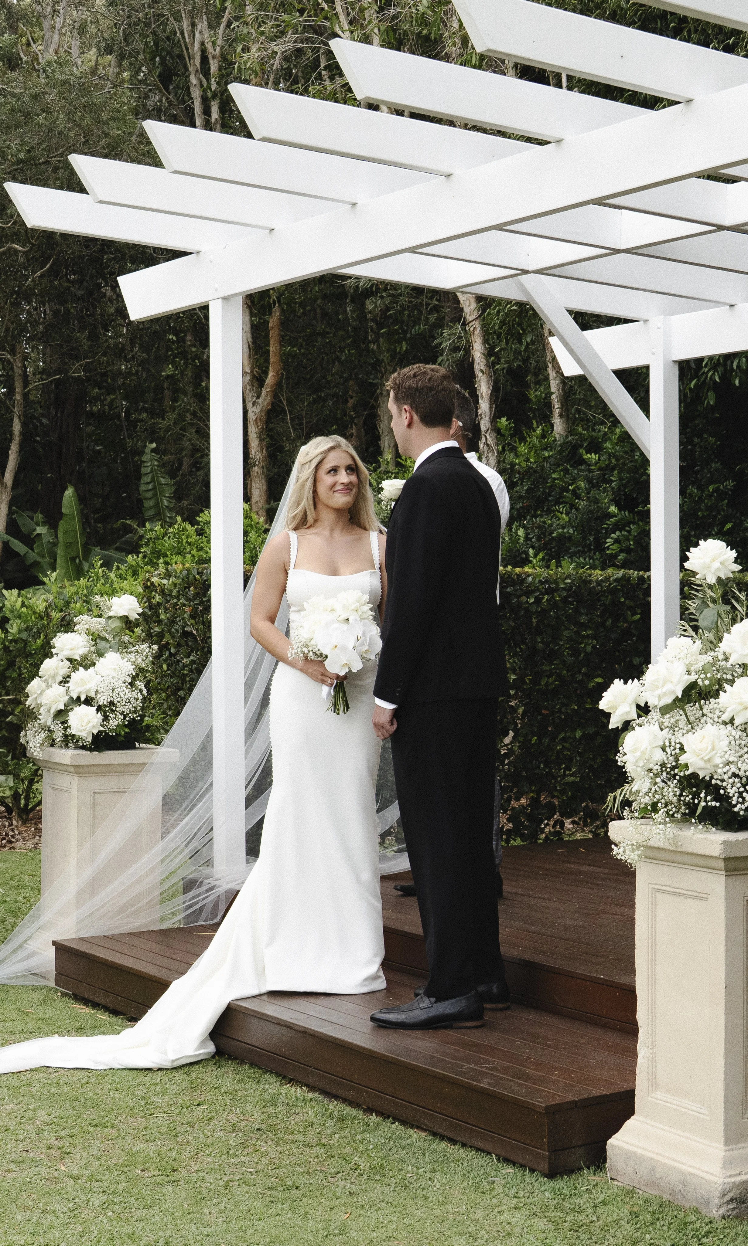 A bride and groom sharing their wedding vows underneath a white pergola surrounded by white floral arrangements in an outdoor garden setting.