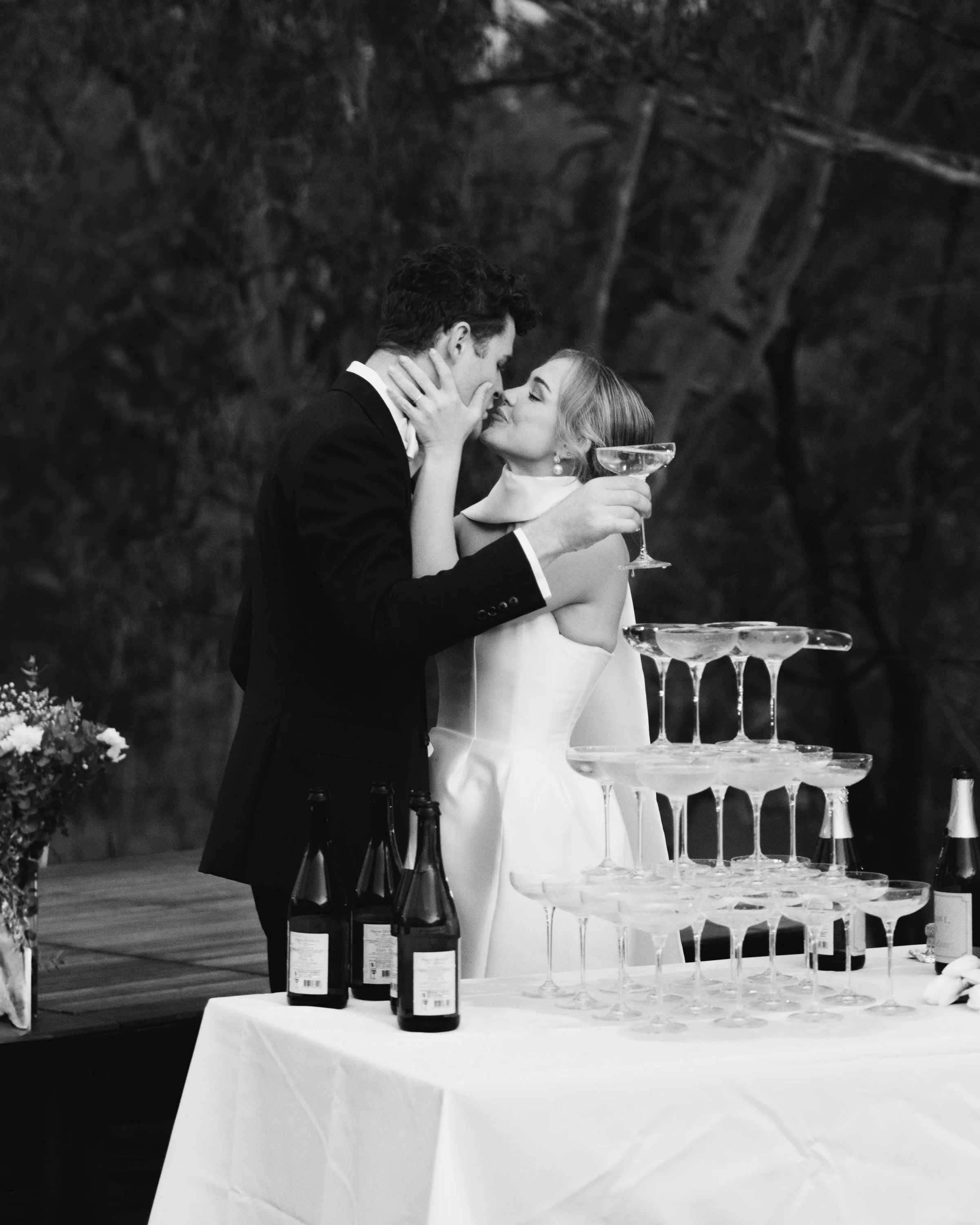 Black and white photo of a couple kissing near a champagne tower and bottles on a table -Elevated Weddings shot on 35mm film