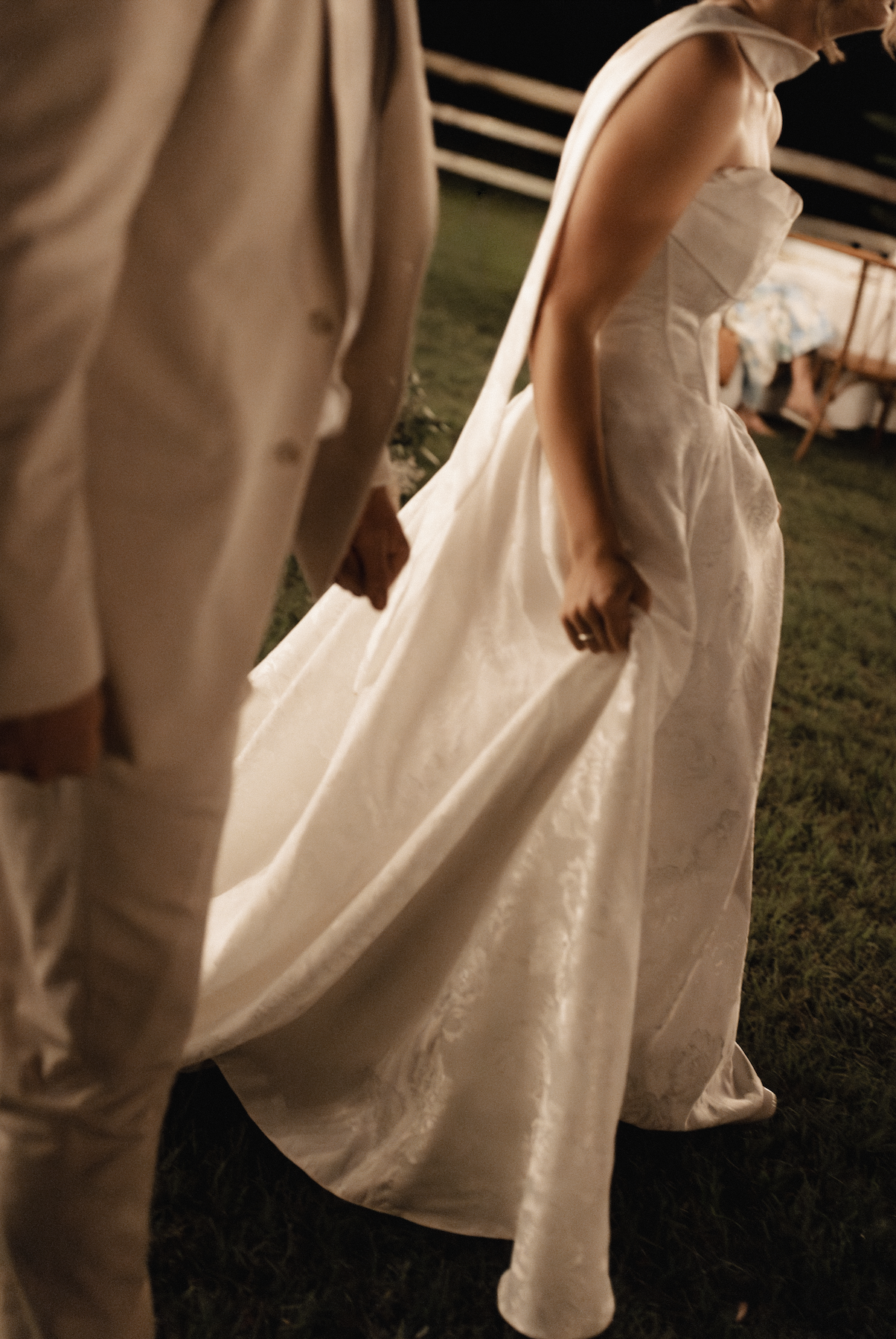 Close-up of a bride holding her wedding dress while standing on grass at night, with a man in a beige suit standing nearby and a table with a patterned tablecloth in the background.