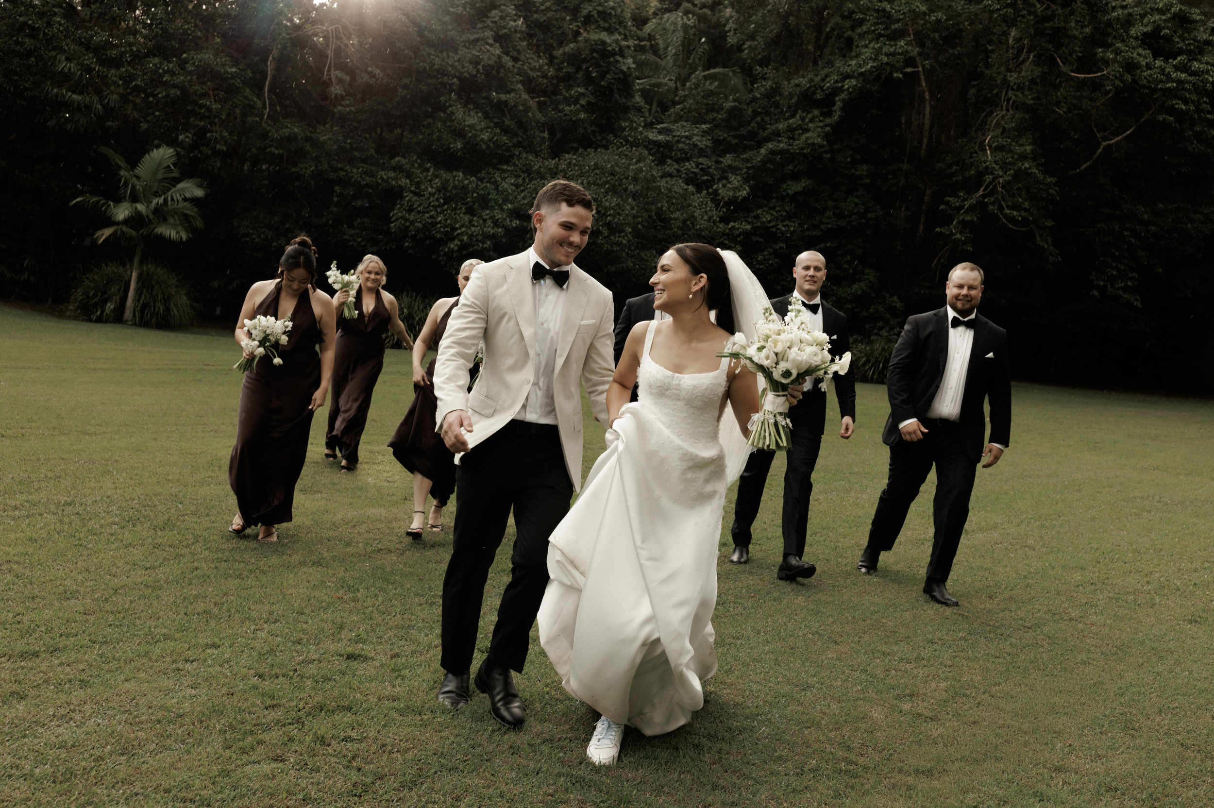 A wedding party walking on a grassy lawn in a wooded area, with the bride and groom smiling at each other in the center, surrounded by bridesmaids and groomsmen dressed in formal attire.