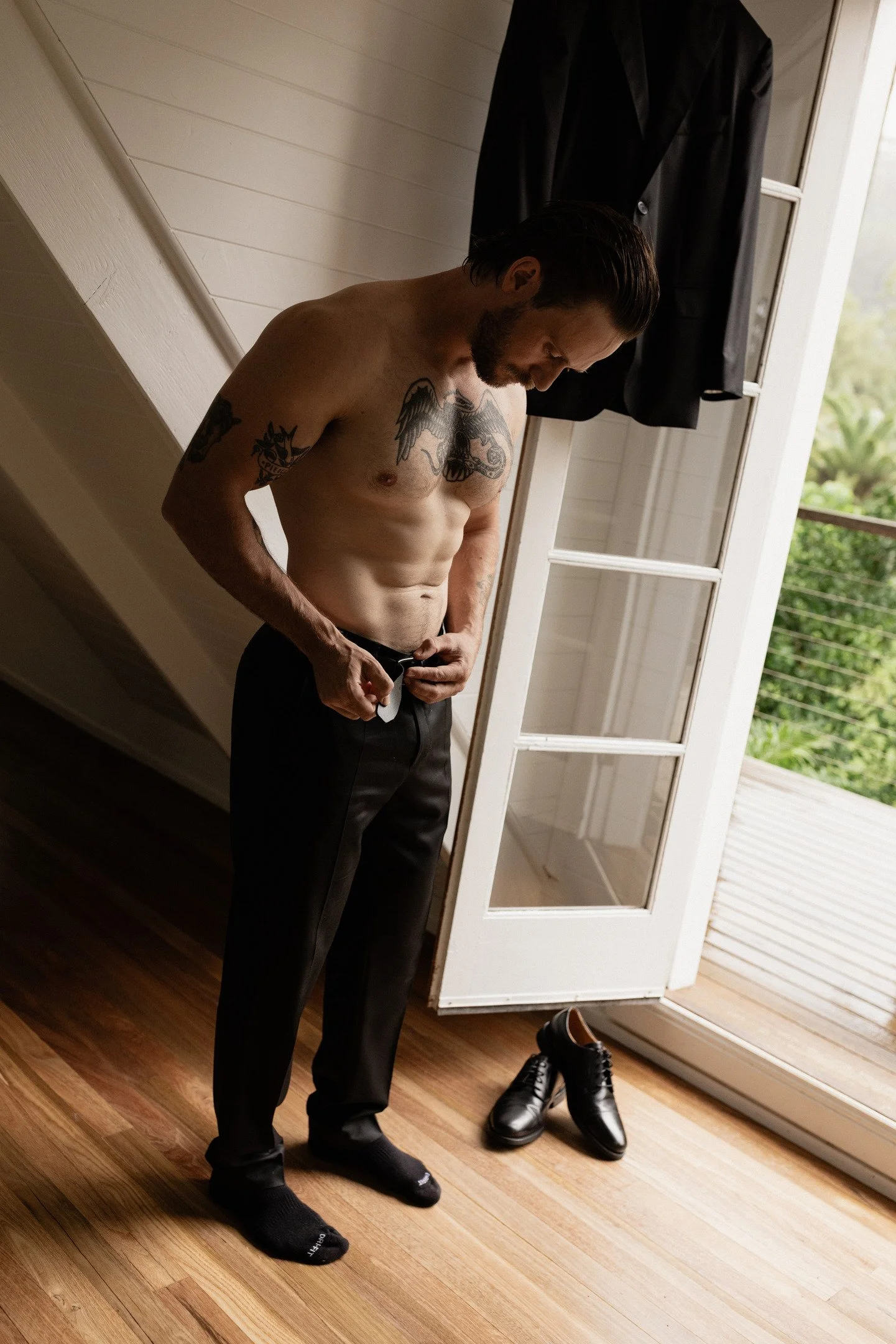 A shirtless man with tattoos is standing on a wooden floor, adjusting his pants. He is wearing black socks and shoes nearby. A black jacket hangs on a hanger behind him in front of a glass door leading to a balcony with greenery outside.