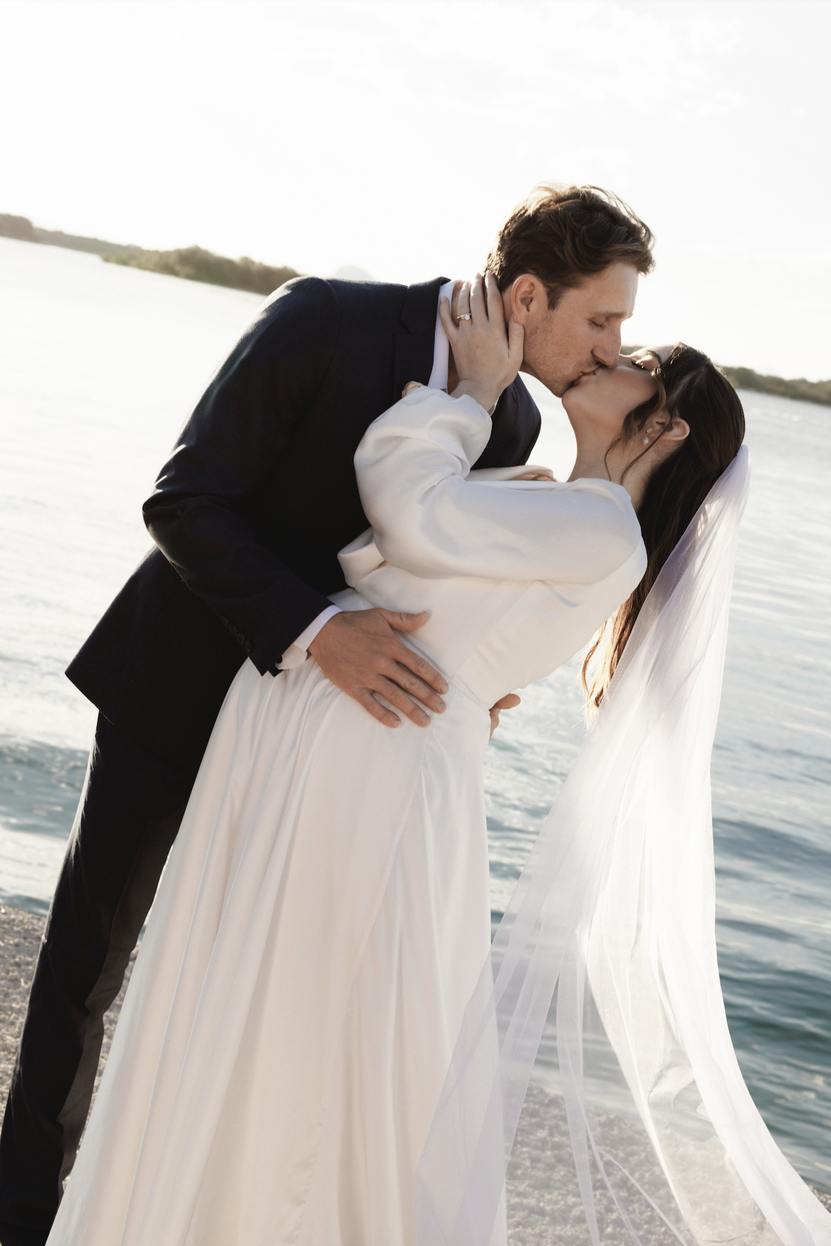 A bride and groom sharing a kiss by the water during sunset, with the bride wearing a white dress and veil, and the groom in a dark suit.