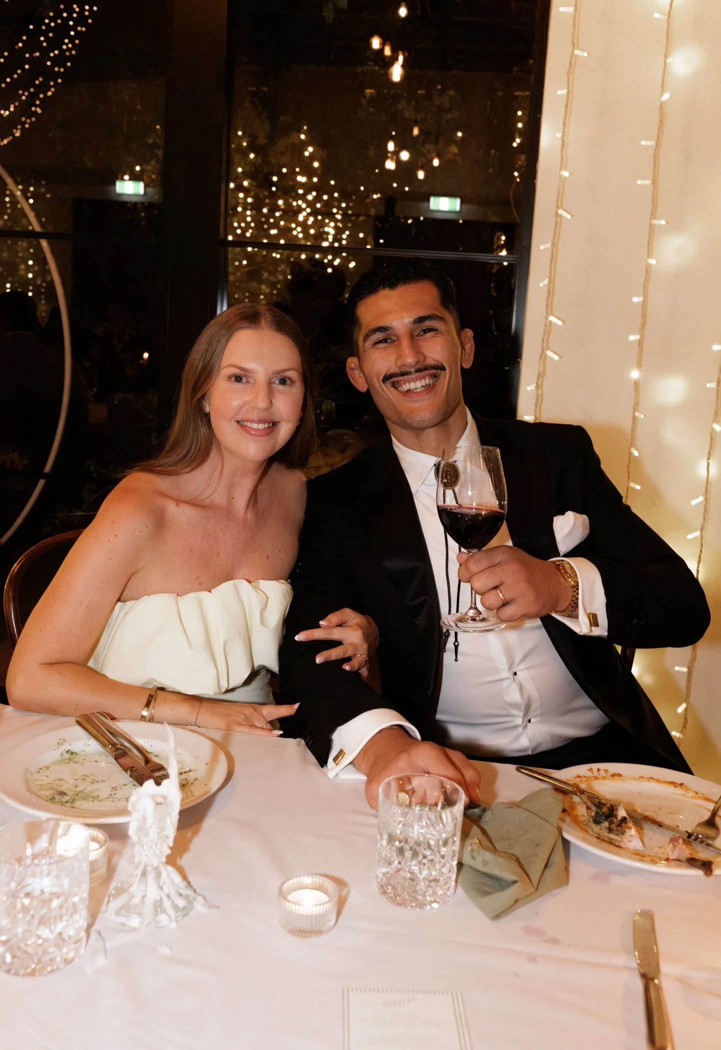 A man and a woman are sitting at a dinner table with empty plates and drinks, smiling at the camera. The man is holding a glass of red wine, wearing a tuxedo with a bow tie and a gold watch. The woman is wearing a strapless white dress with ruffles, 
