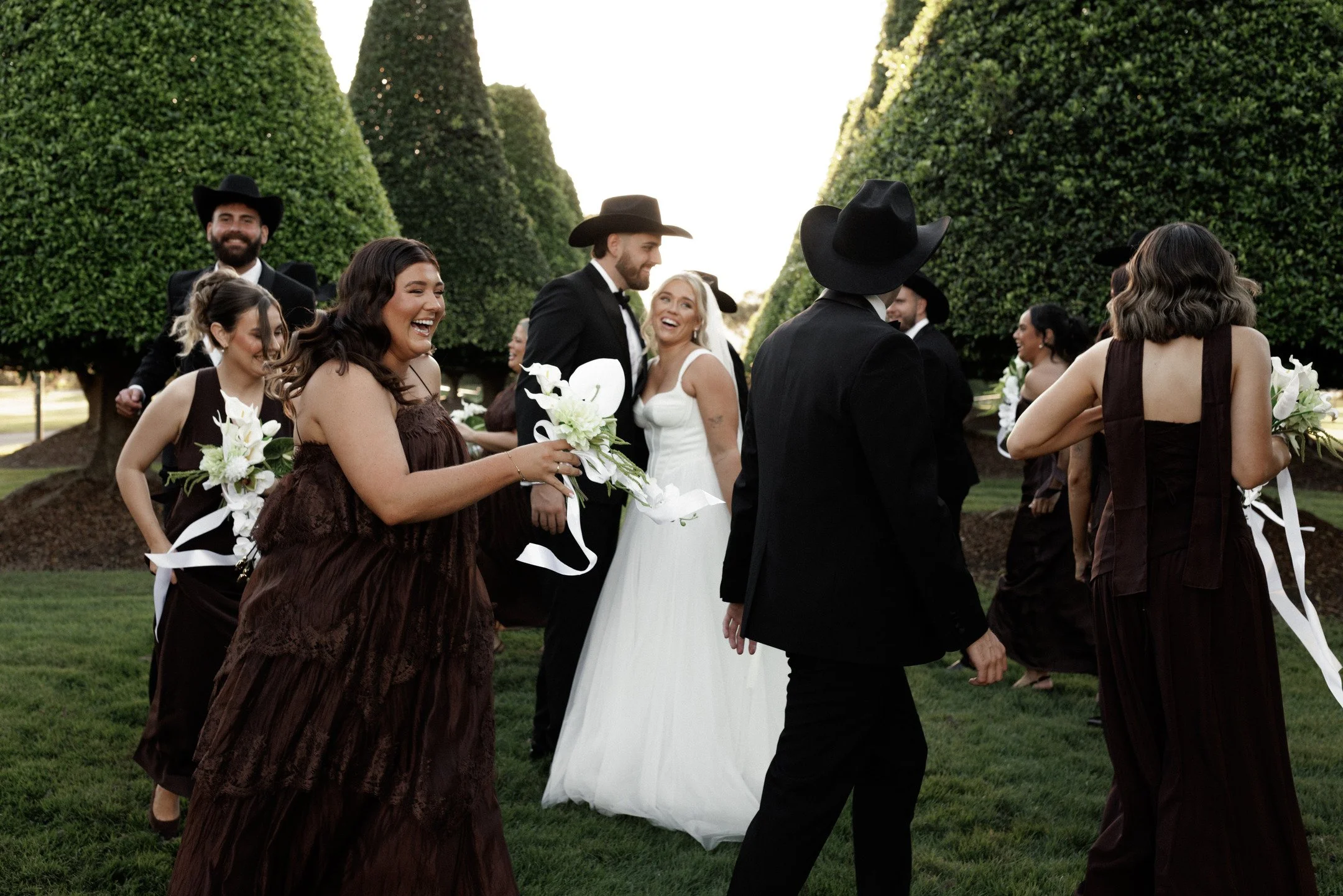 A wedding party is walking outdoors between tall, neatly trimmed hedges. The group includes men in black suits and wide-brimmed hats, women in elegant brown and black dresses, and a bride in a white wedding dress holding a bouquet. Everyone appears h