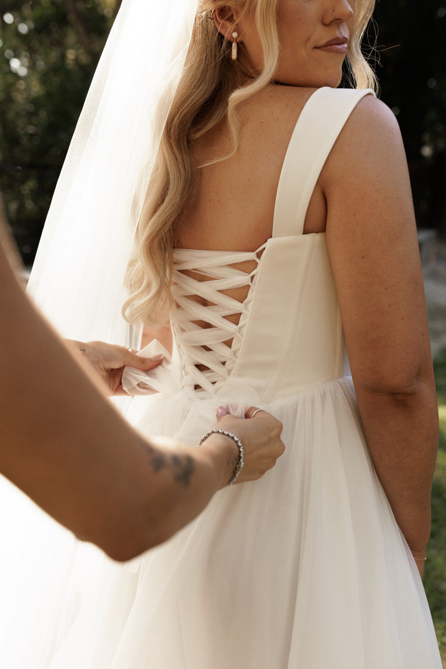 Close-up of a bride wearing a white dress with lace-up back, during a wedding ceremony. Someone helps tie the dress, with fingers visible near the waist. The bride has long, wavy blonde hair, earrings, and makeup.
