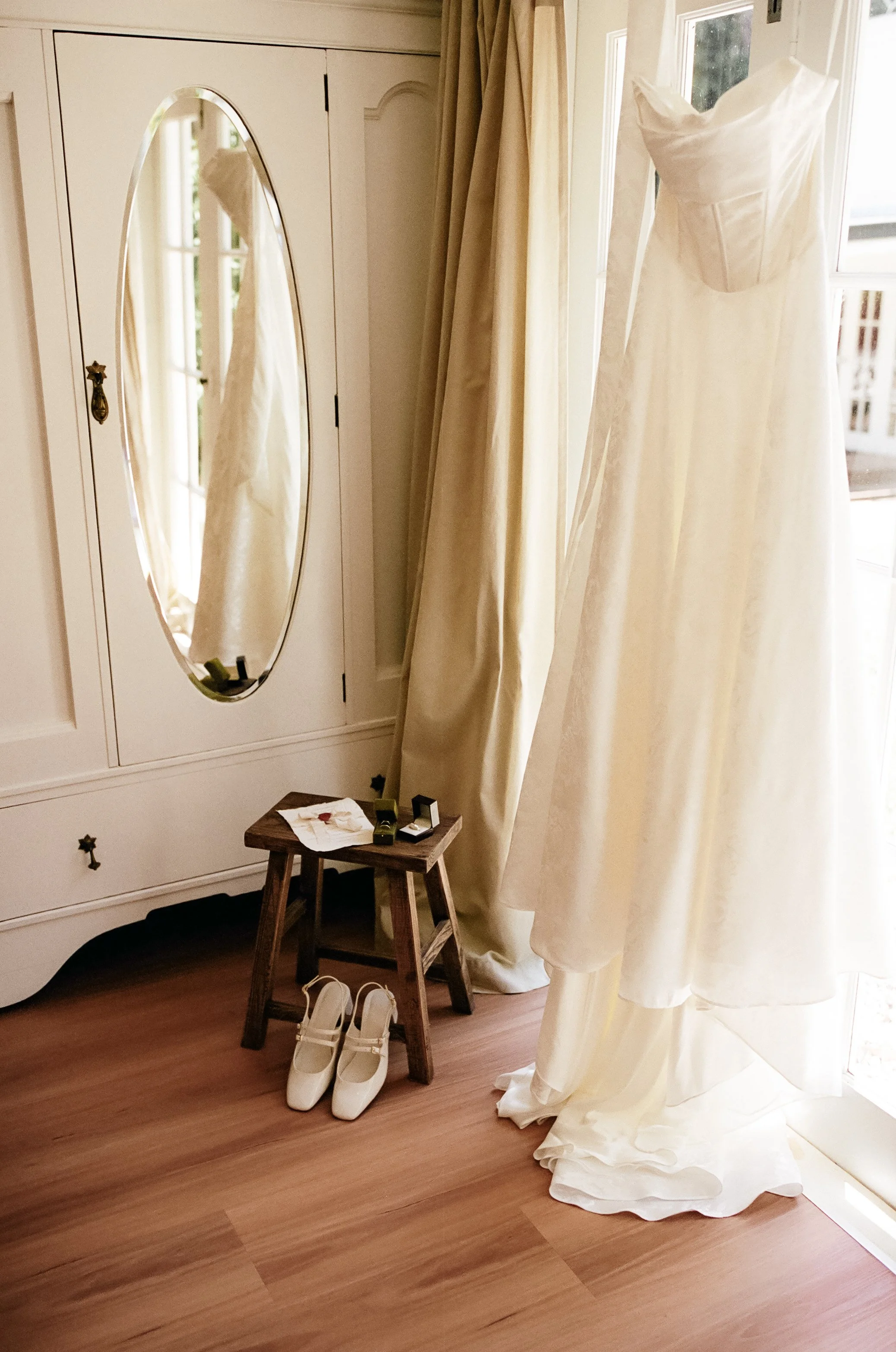 A white wedding dress hanging near a window, with a pair of white shoes below it, a small wooden stool, and a mirror reflecting part of the dress.