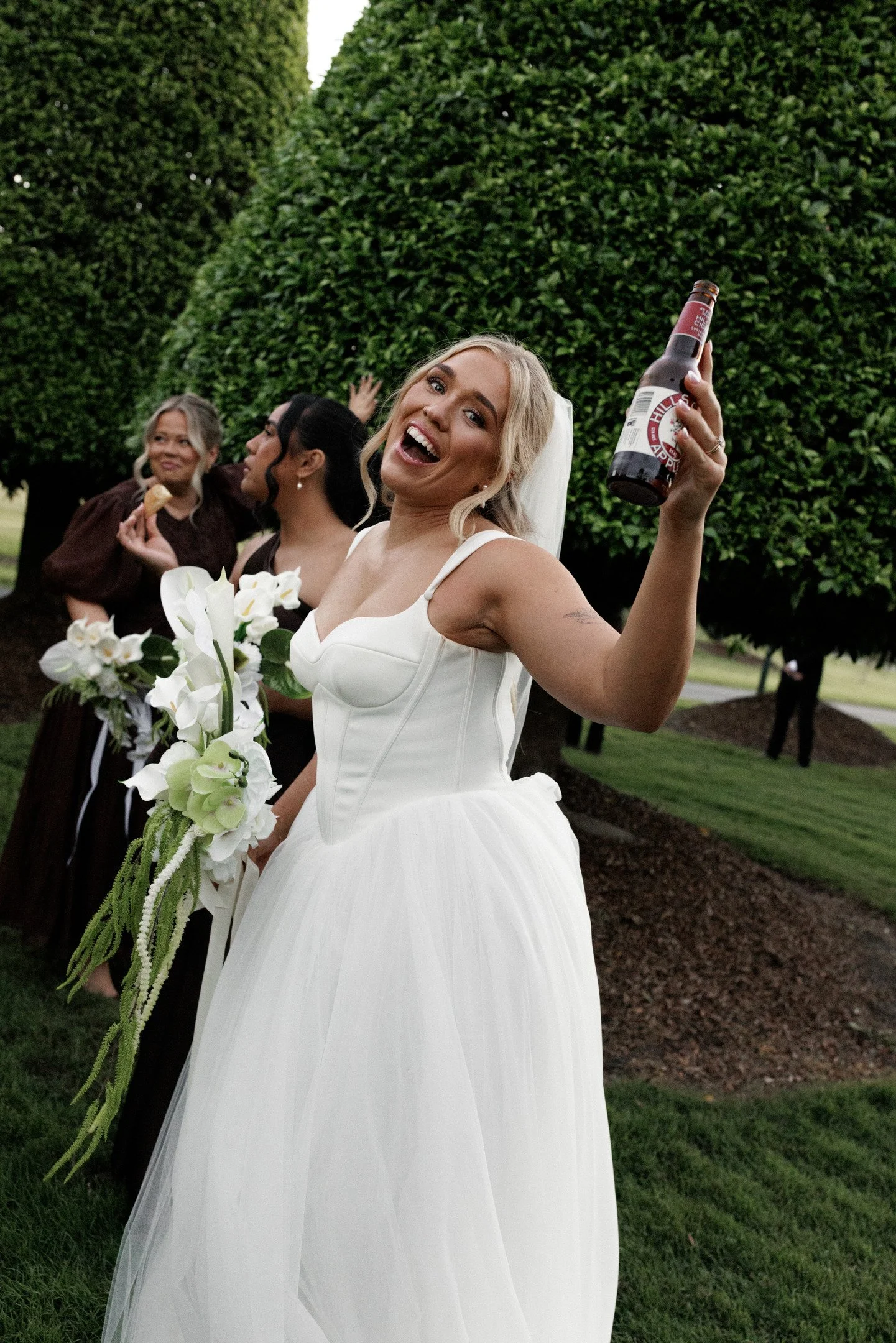 A smiling bride in a white wedding gown holding a bouquet of white flowers in one hand and a beer bottle in the other, standing outdoors with green trees in the background.