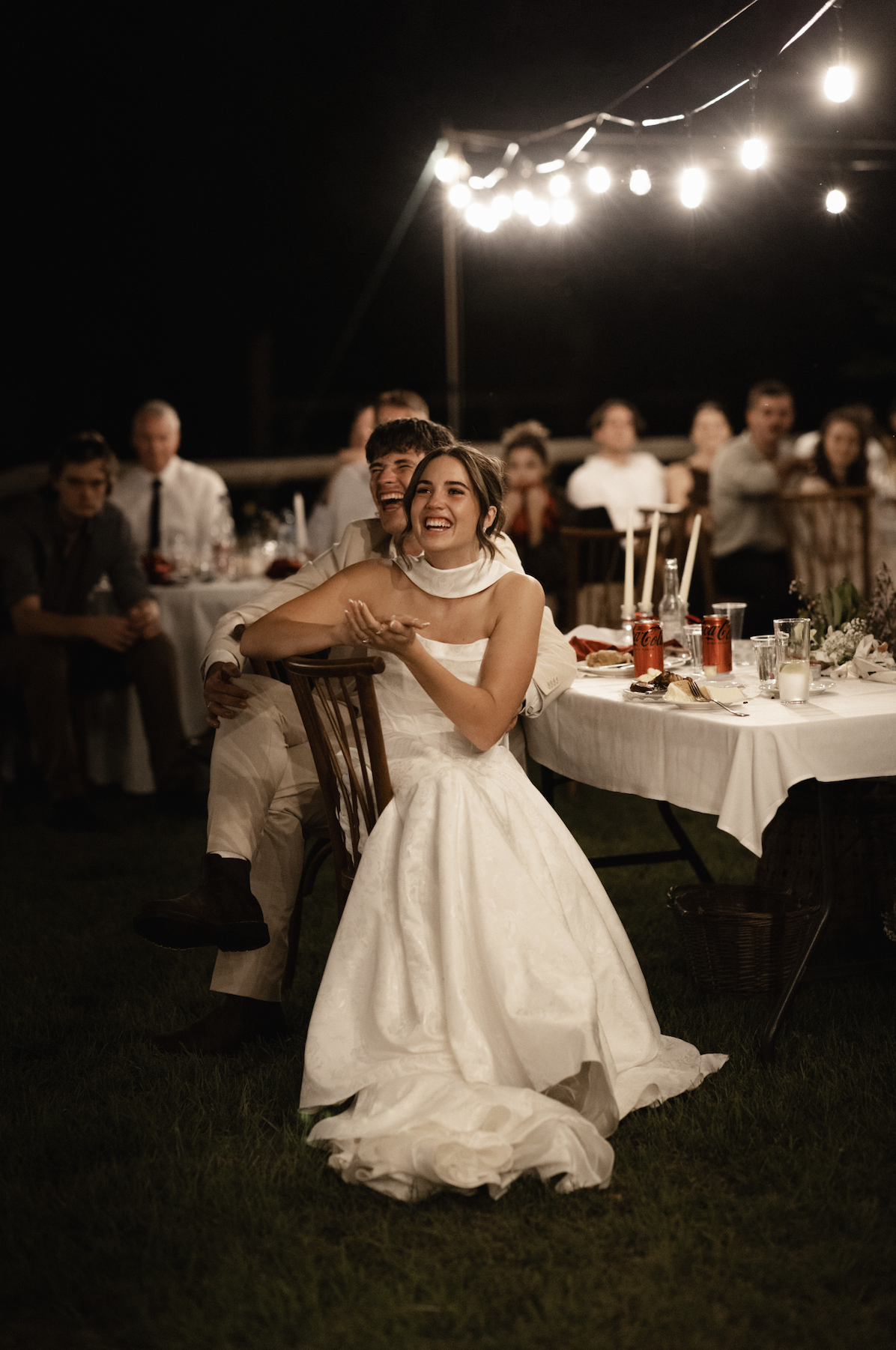 A bride and groom sitting together at their wedding reception, smiling and laughing under string lights at night, with guests seated at tables around them.