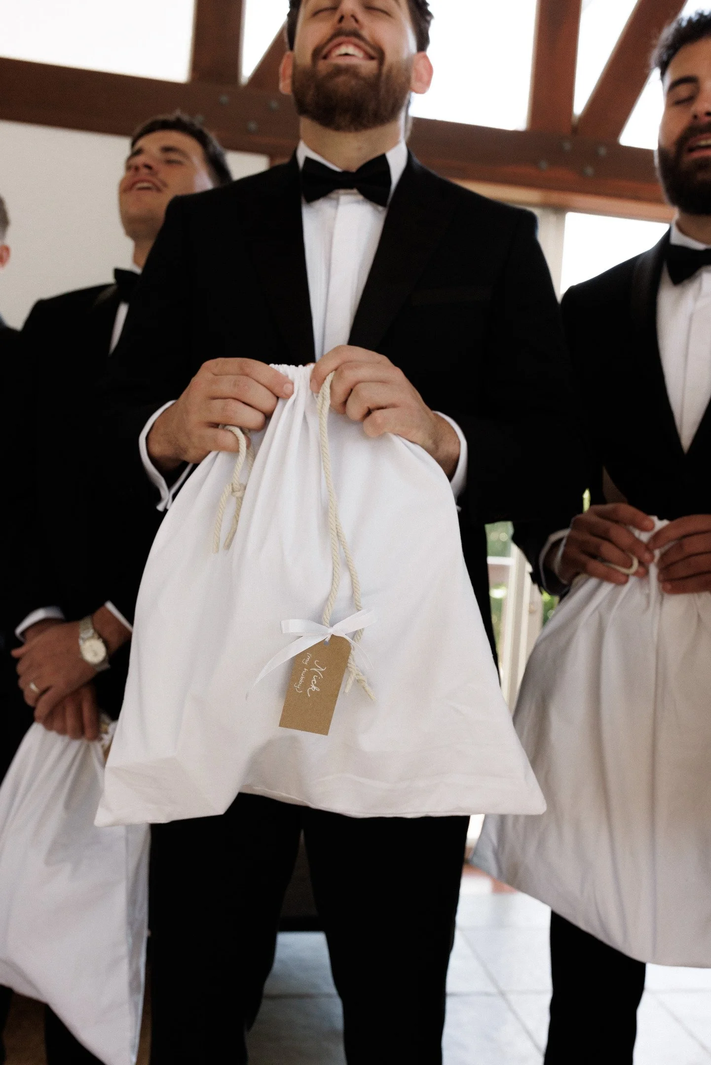 A group of men dressed in tuxedos holding white gift bags, standing indoors with a wooden ceiling, smiling and enjoying the moment.