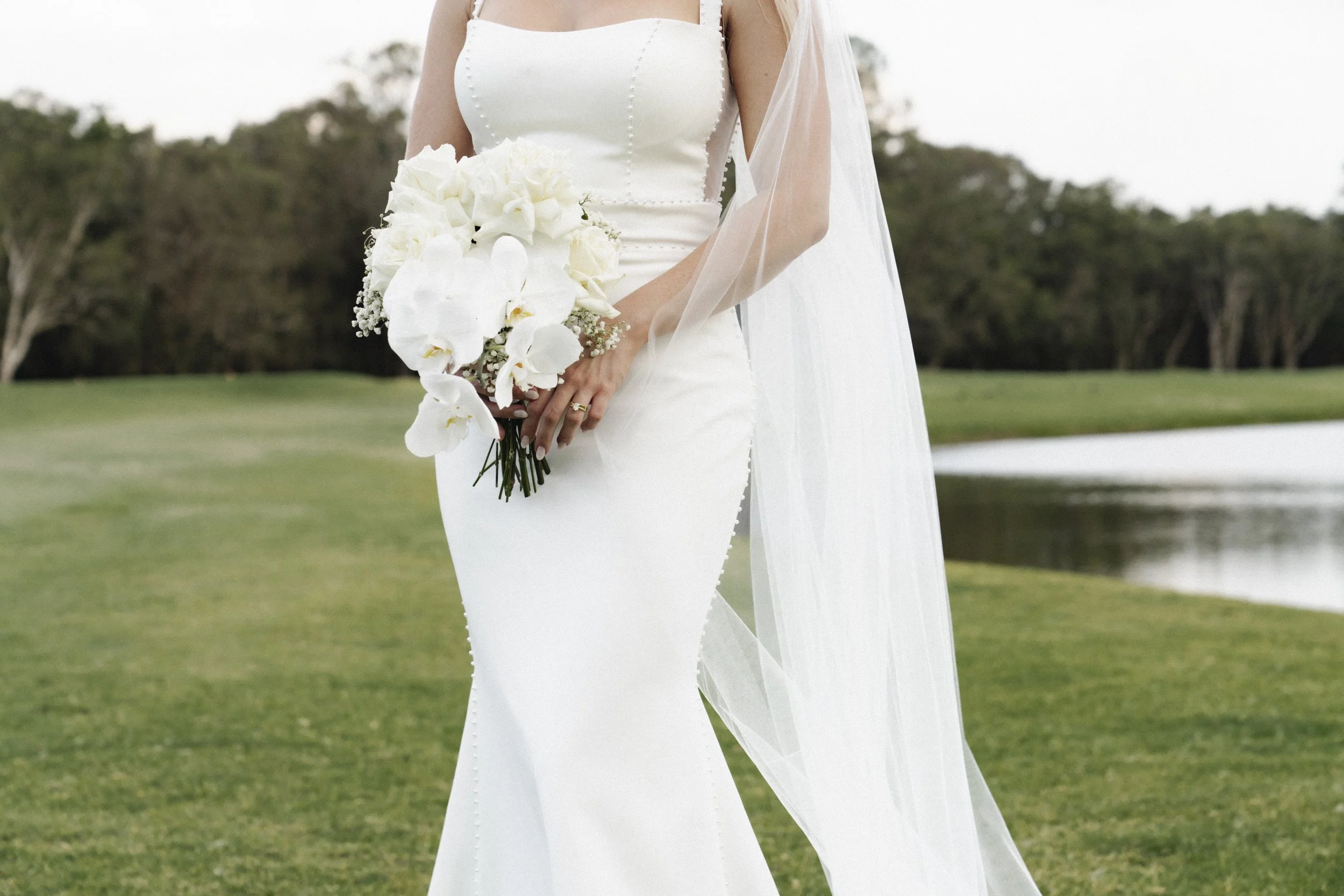 Bride in white dress holding a bouquet of white flowers, standing outdoors near a pond - Elevated Weddings