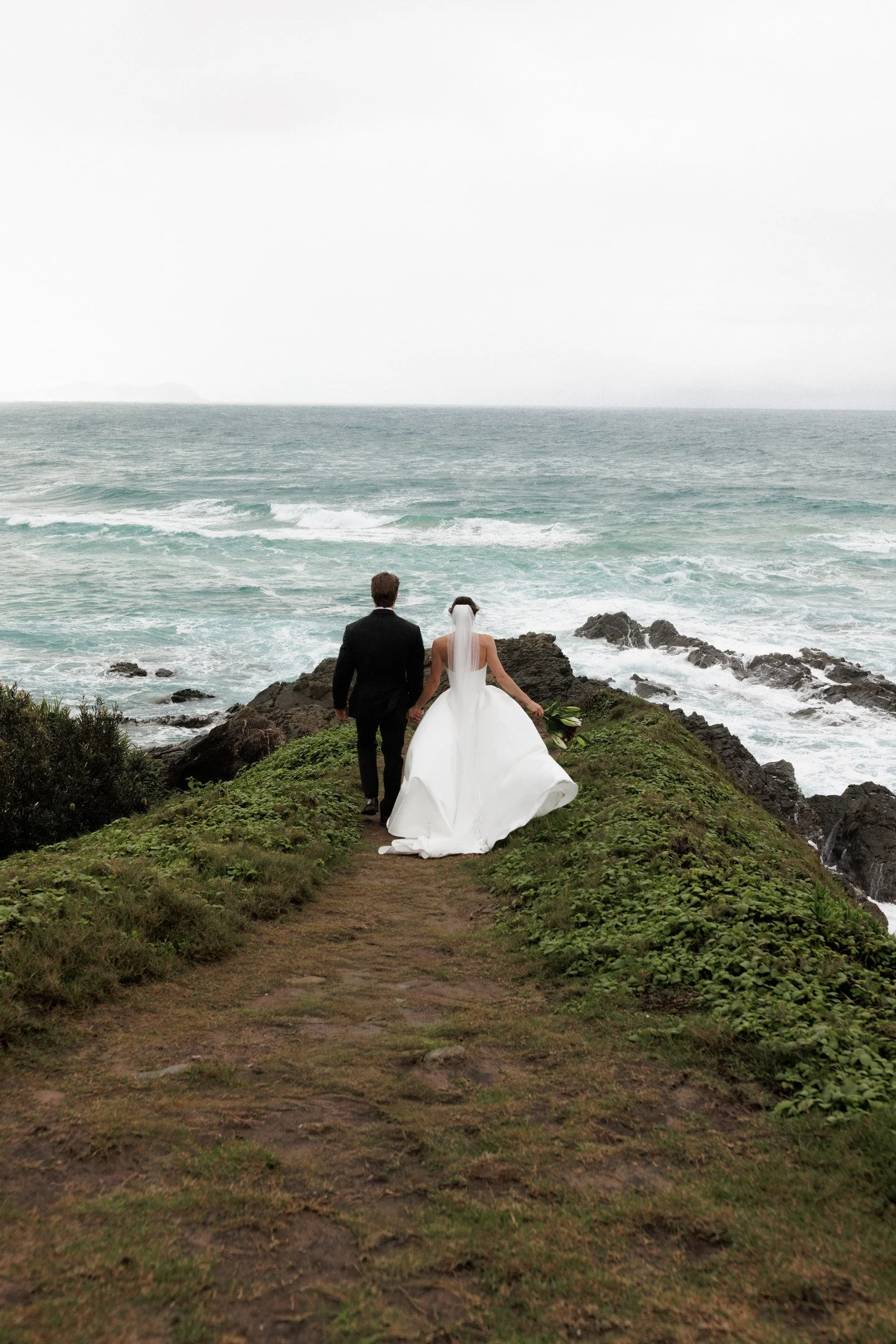 A bride and groom holding hands, walking on a grassy path along the coast with rocky shores and ocean waves, under a cloudy sky.