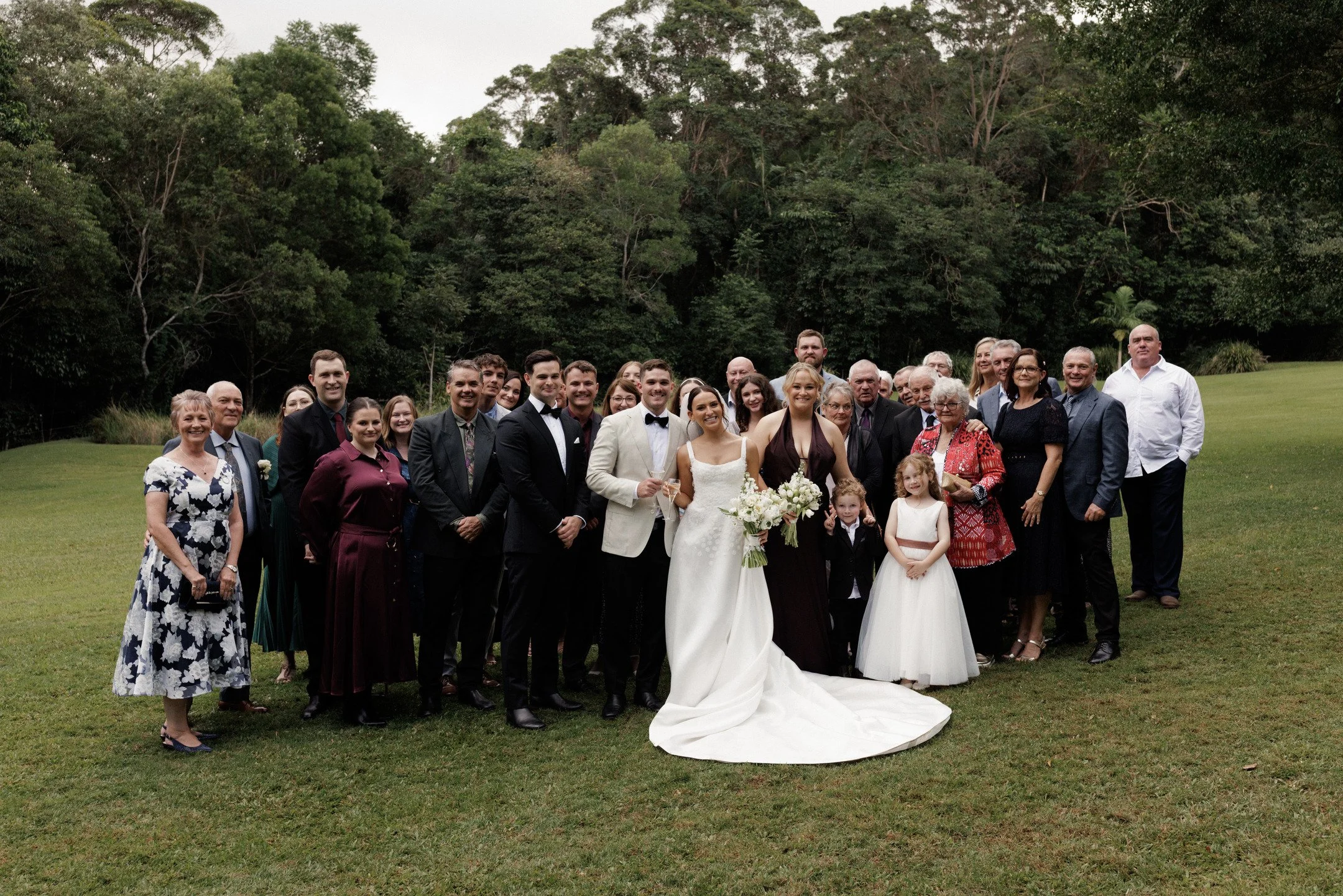 Group of wedding guests and the bride and groom standing on a grassy field with trees in the background, smiling and posing for a photo.