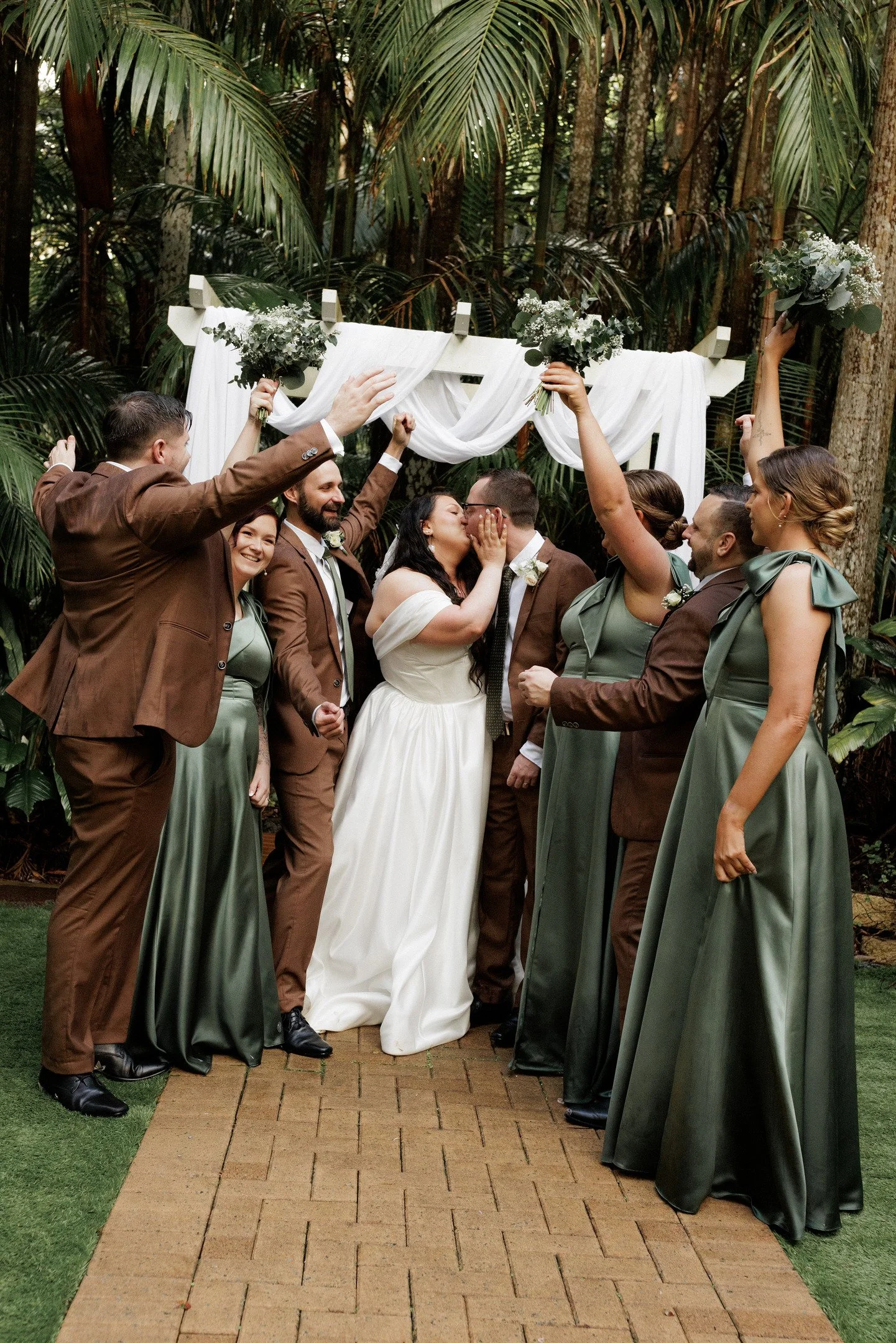 A wedding ceremony in an outdoor garden setting with a couple kissing as the officiant and wedding party celebrate around them, floral bouquets are held up in the air, and the background features lush greenery and trees.
