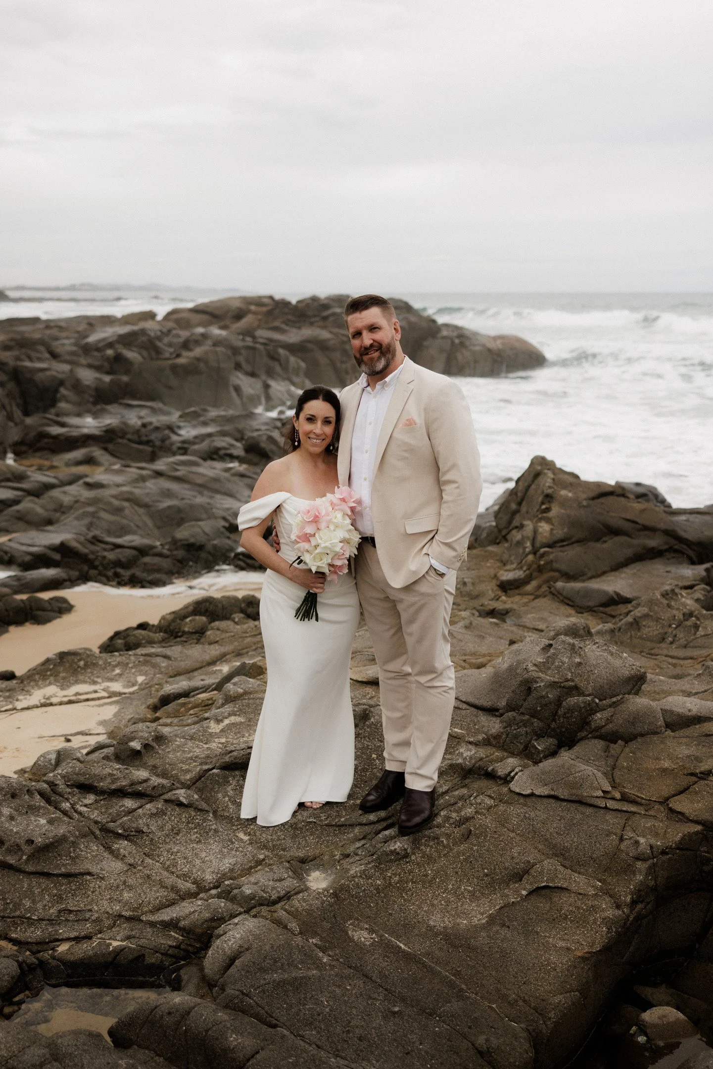 A newlywed couple stands on rocky beach near the ocean, dressed in wedding attire. The bride wears a white dress and holds a bouquet of pink and white flowers, while the groom wears a cream-colored suit with a white shirt. Overcast sky and waves crashing against rocks behind them.