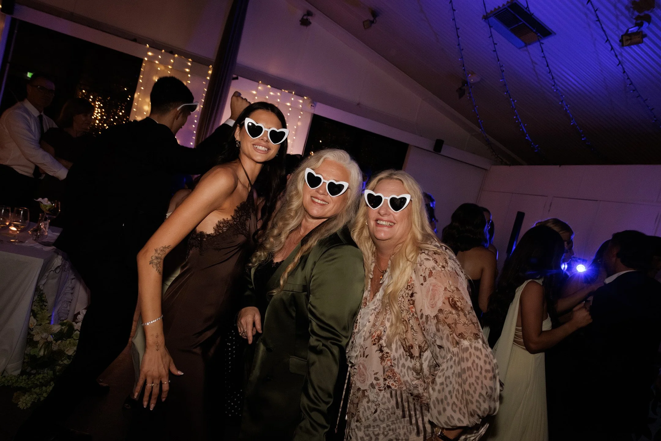 Three women wearing heart-shaped sunglasses smiling at a party or celebration, with a background of other guests and decorative string lights.