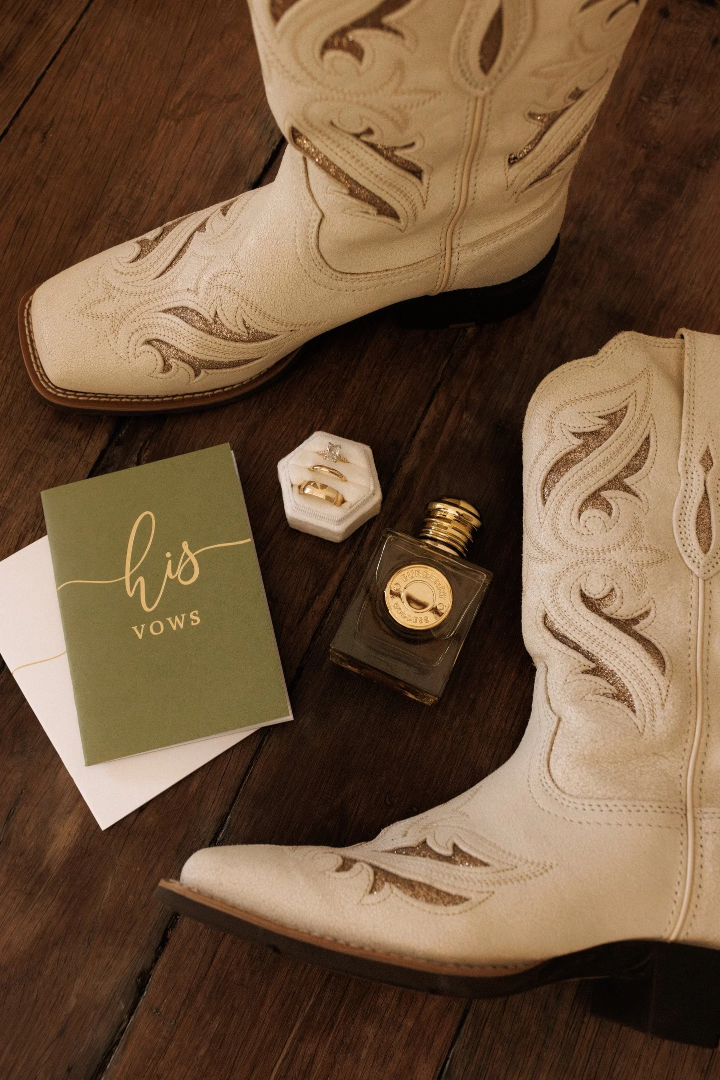A pair of cream-colored cowboy boots with decorative stitching on the leather, on a wooden floor. Near the boots, there are wedding rings in a small white box, a perfume bottle, and a green card with the words "his vows" written on it.