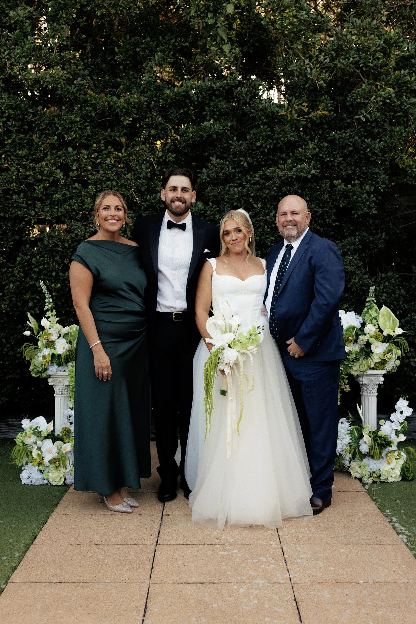 A group of four people, two women and two men, standing together outdoors in front of lush green bushes. The woman on the left is wearing a dark green dress, and the woman in the center is wearing a white wedding gown and holding a bouquet. The man n