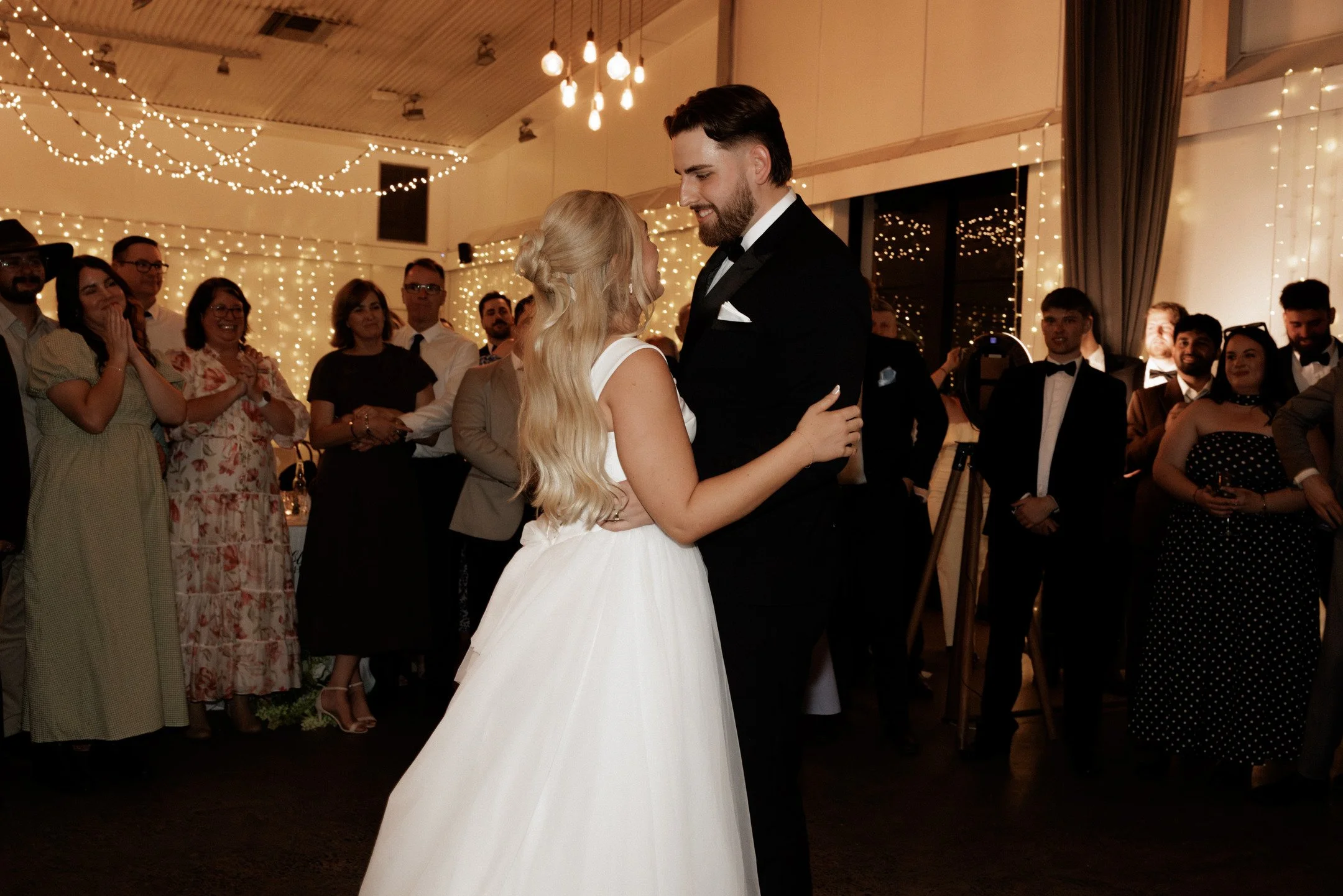 A bride and groom dancing during their wedding reception, surrounded by smiling guests under hanging string lights.