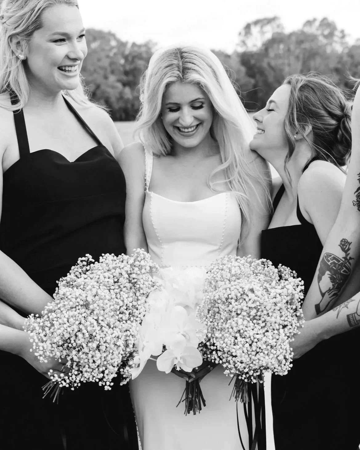 A group of women, with one in a white dress holding a bouquet of flowers, smiling and laughing outdoors.