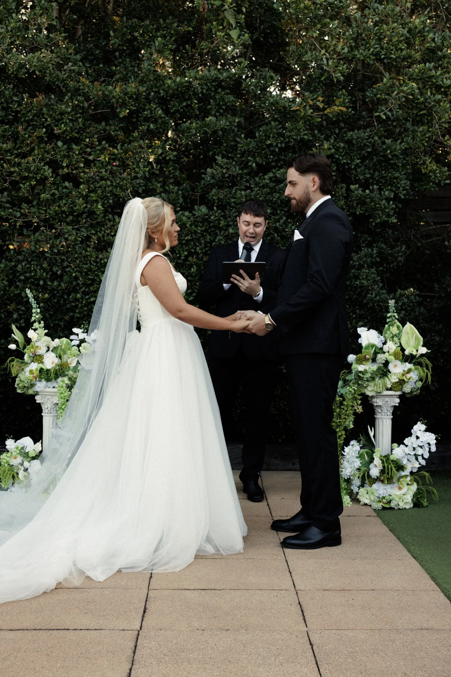 A bride and groom standing hand in hand exchanging vows during a wedding ceremony outdoors, with an officiant reading from a book and decorated floral arrangements nearby.