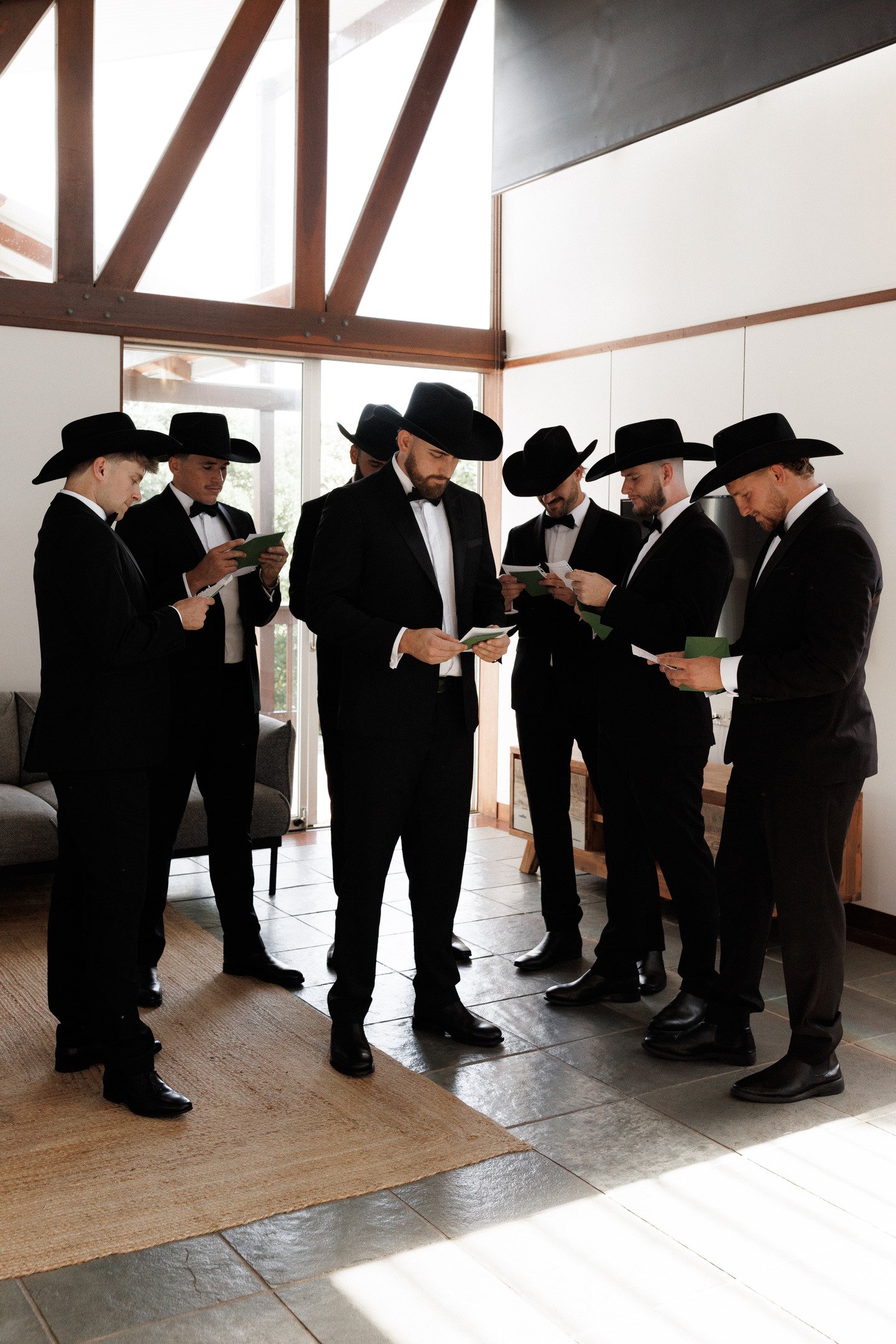 Seven men dressed in black tuxedos and cowboy hats standing together indoors, reading papers.