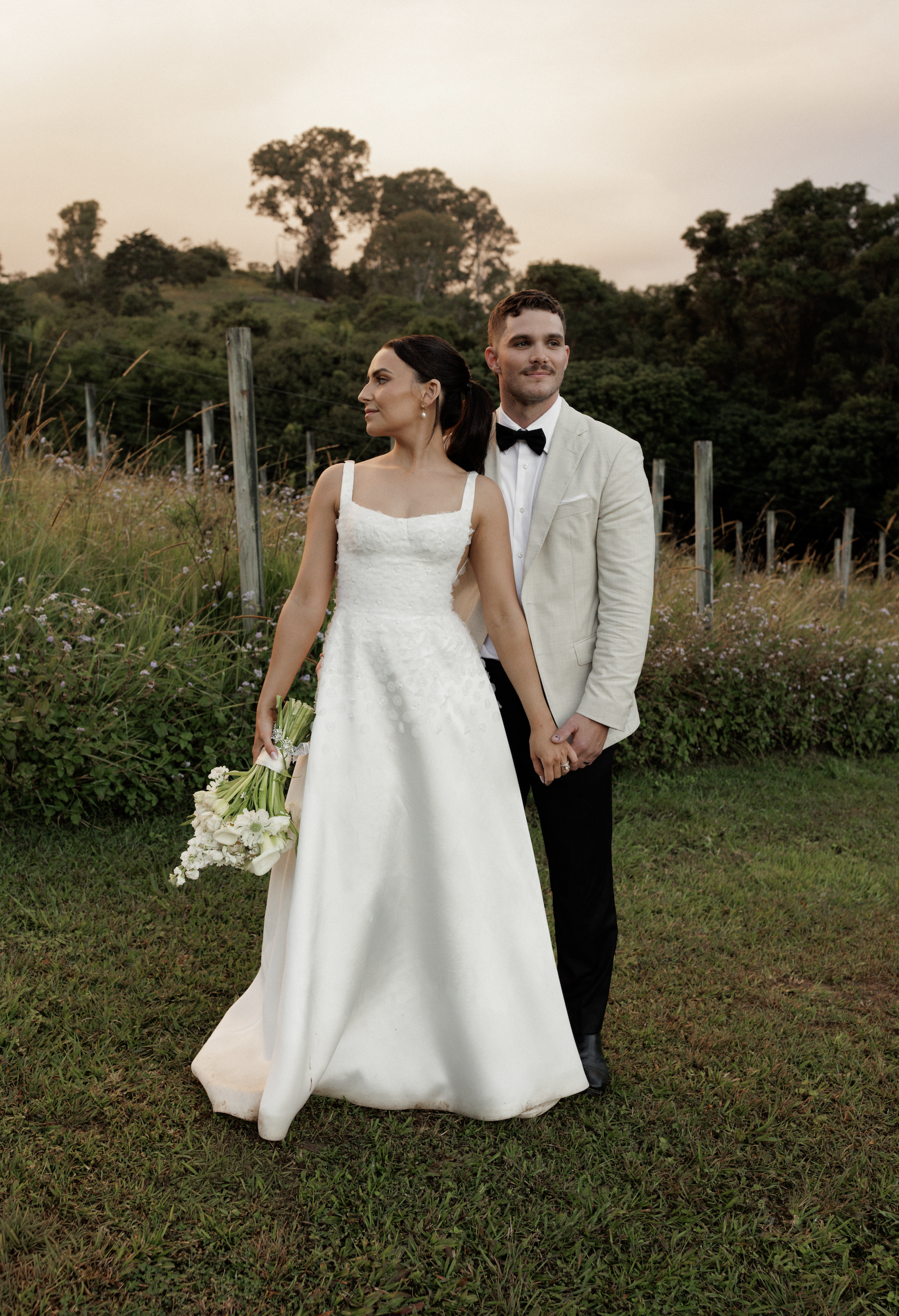 A bride and groom standing outdoors on a grassy field with trees and a fence in the background during sunset, holding hands and dressed in wedding attire.